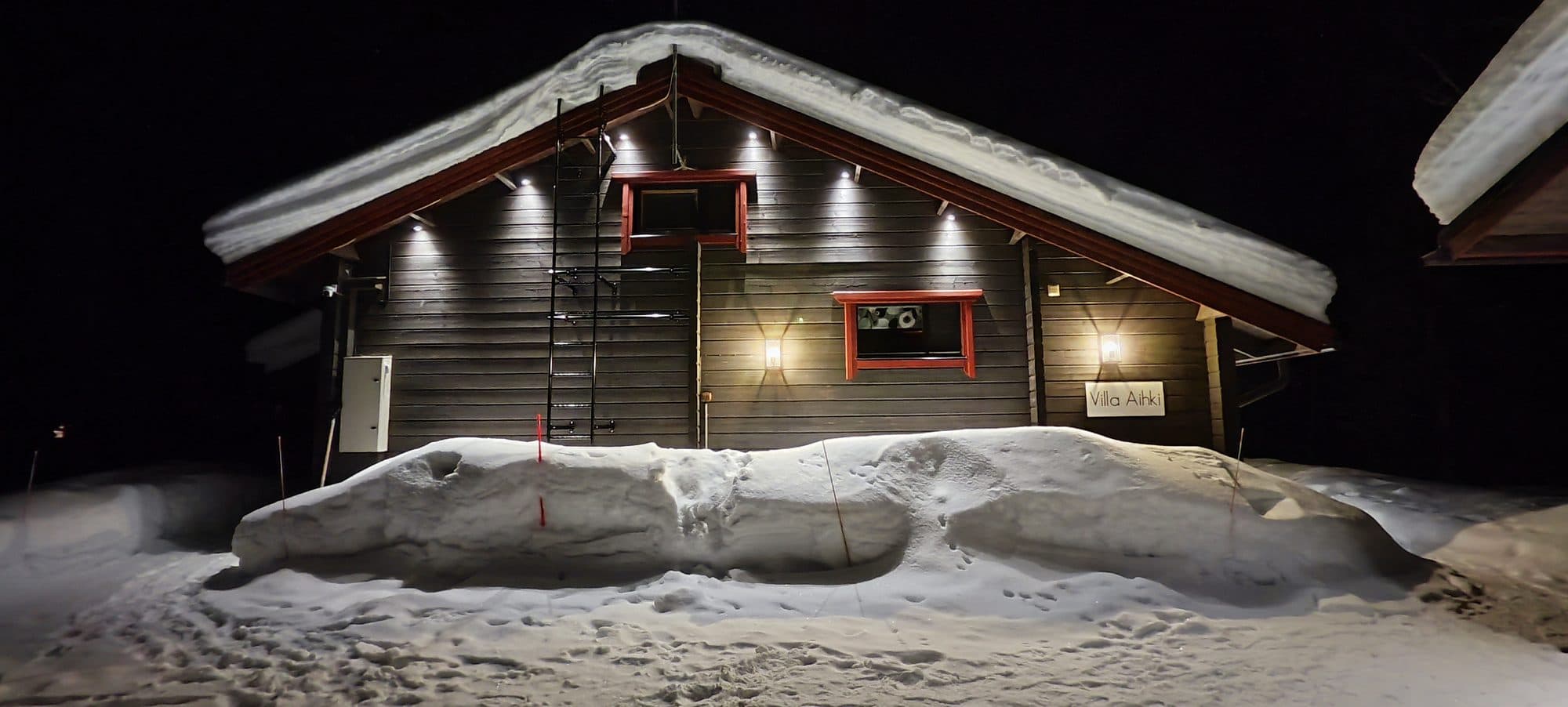 Nächtliche Ansicht einer beleuchteten Holzhütte mit schneebedecktem Dach und einer hohen Schneewehe im Vorgarten.