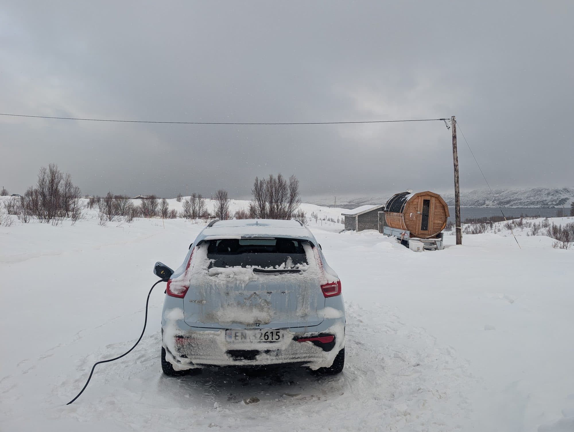 Ein weißes Elektroauto lädt im tiefen Schnee vor einer traditionellen runden Holzsauna.