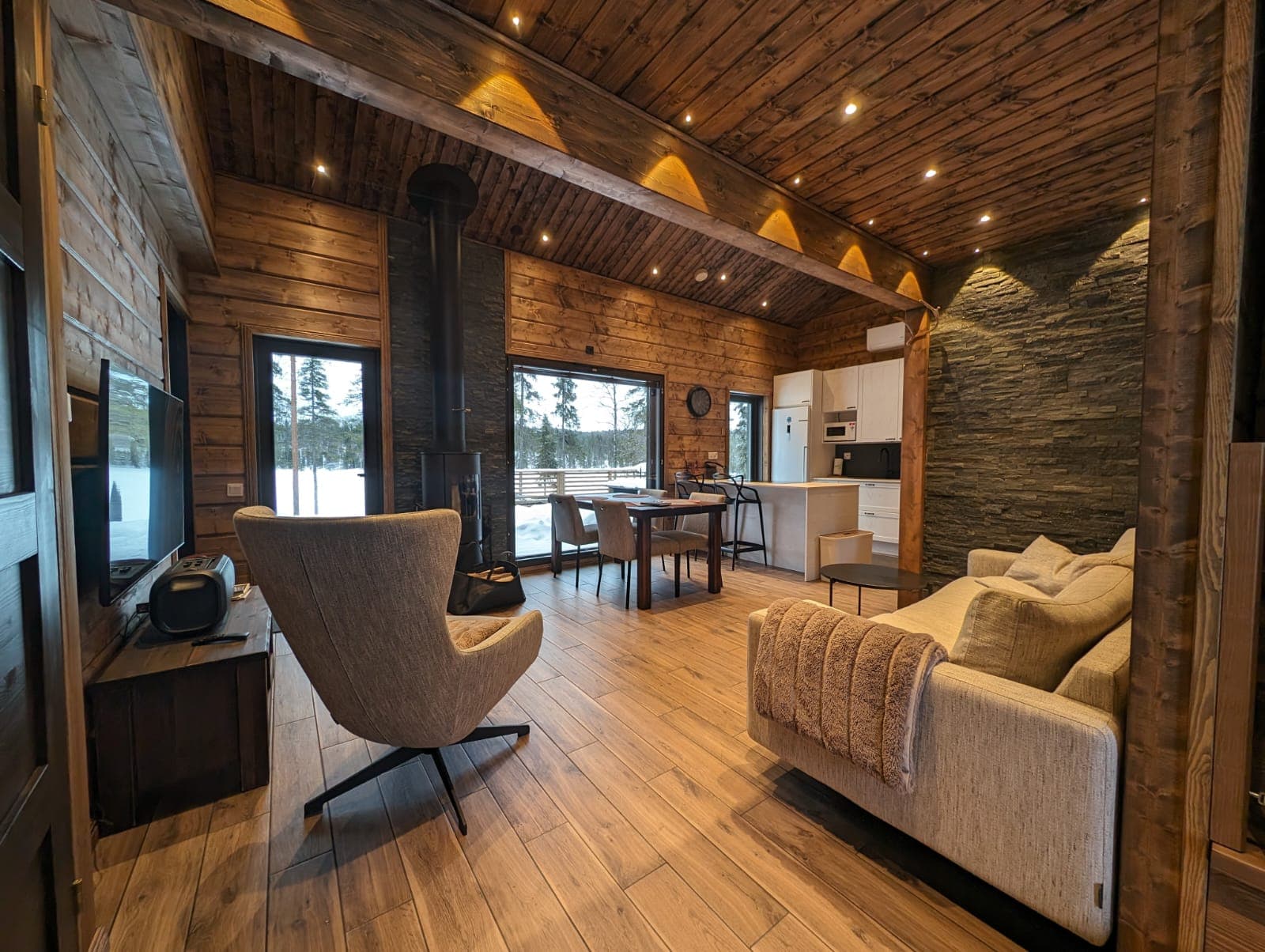 Offener Wohn-Ess-Küchenbereich einer Ferienhütte in Kuusamo mit Kaminofen, Holzverkleidung und Panoramafenstern zur verschneiten Waldlandschaft.