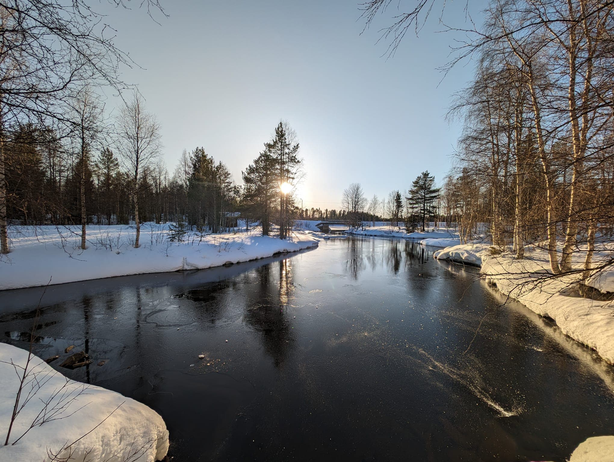 Die tiefstehende Wintersonne bricht sich golden im offenen Wasser eines Flusses bei Kuusamo, umrahmt von schneebedeckten Ufern und kahlen Birken – im Hintergrund eine kleine Brücke.