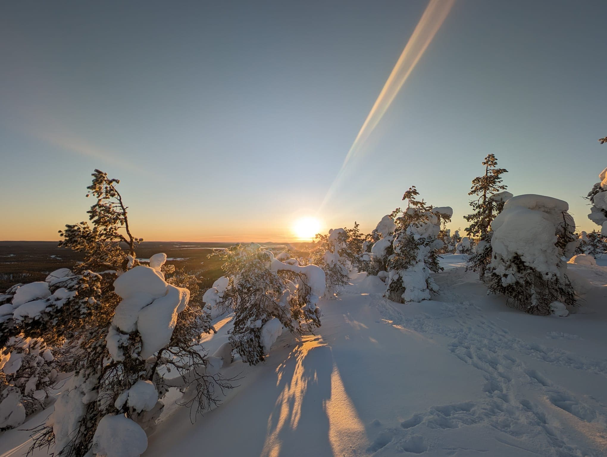 Von einem verschneiten Fjellgipfel mit raureifbedeckten Bäumen blickt man über die endlose Waldlandschaft Kuusamos zur tiefstehenden Sonne am Horizont, Fußspuren ziehen sich durch den Tiefschnee. Der Himmel leuchtet in warmen Orangetönen.