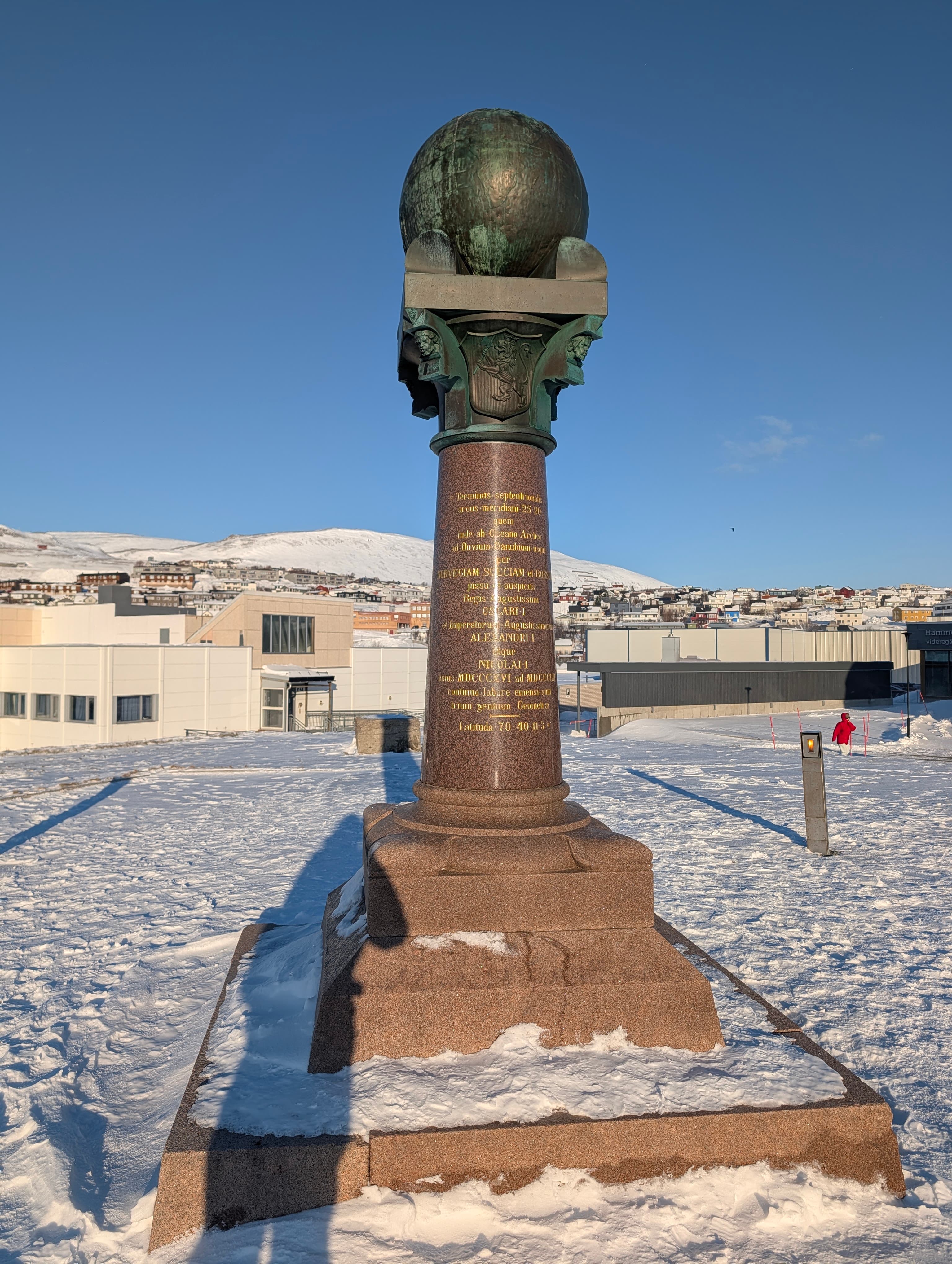 Ein markantes steinernes Denkmal mit Bronzekugel thront über einer verschneiten Stadtlandschaft unter strahlend blauem Himmel. Die Szene vermittelt eine klare, kalte Winteratmosphäre in Nordnorwegen.