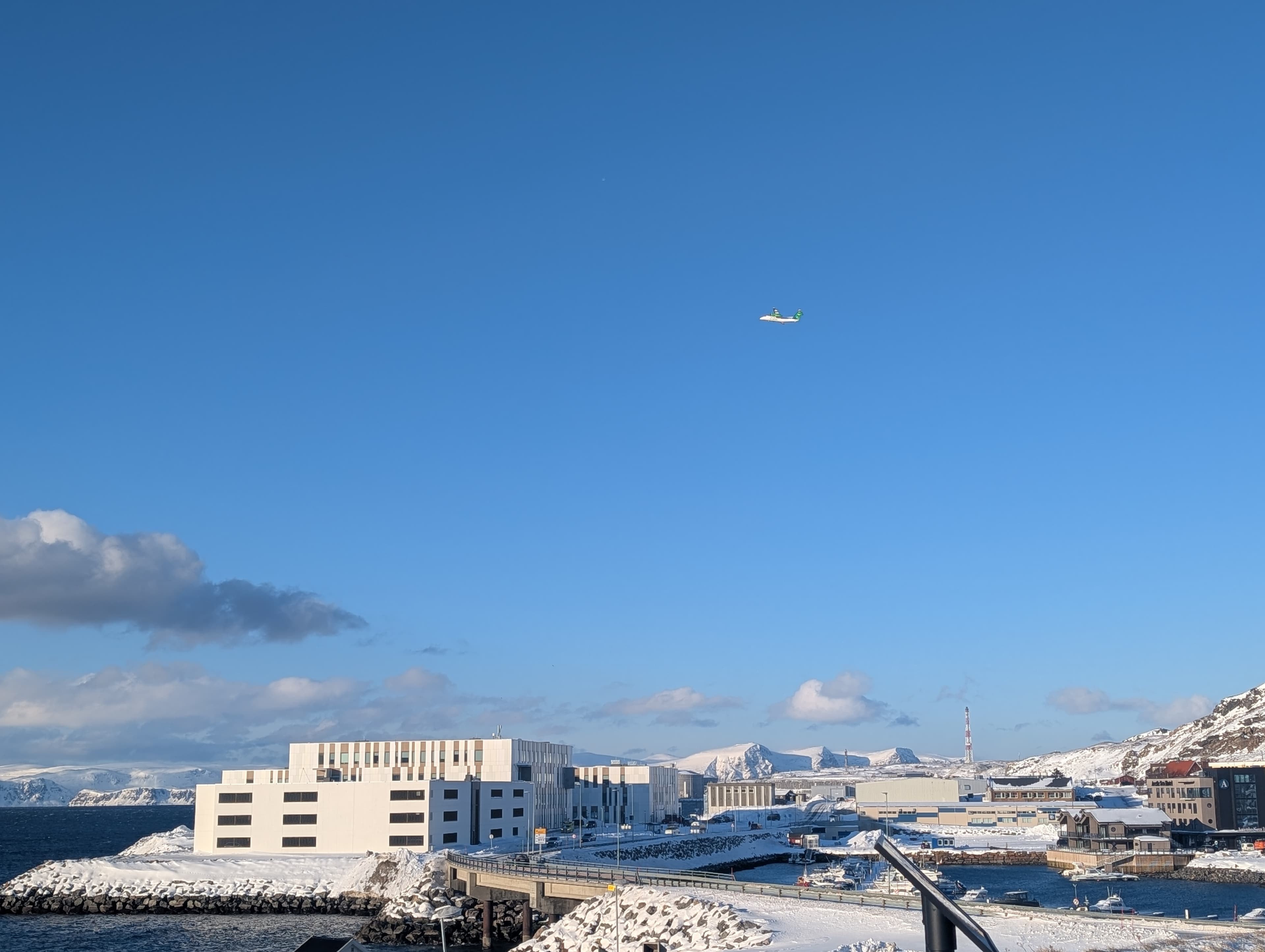 Eine sonnige Winteransicht einer norwegischen Hafenstadt mit moderner Architektur und schneebedeckten Bergen im Hintergrund, während ein Flugzeug am blauen Himmel vorbeizieht. Die Szene strahlt kühle Klarheit aus und zeigt den Alltag in Nordnorwegen.