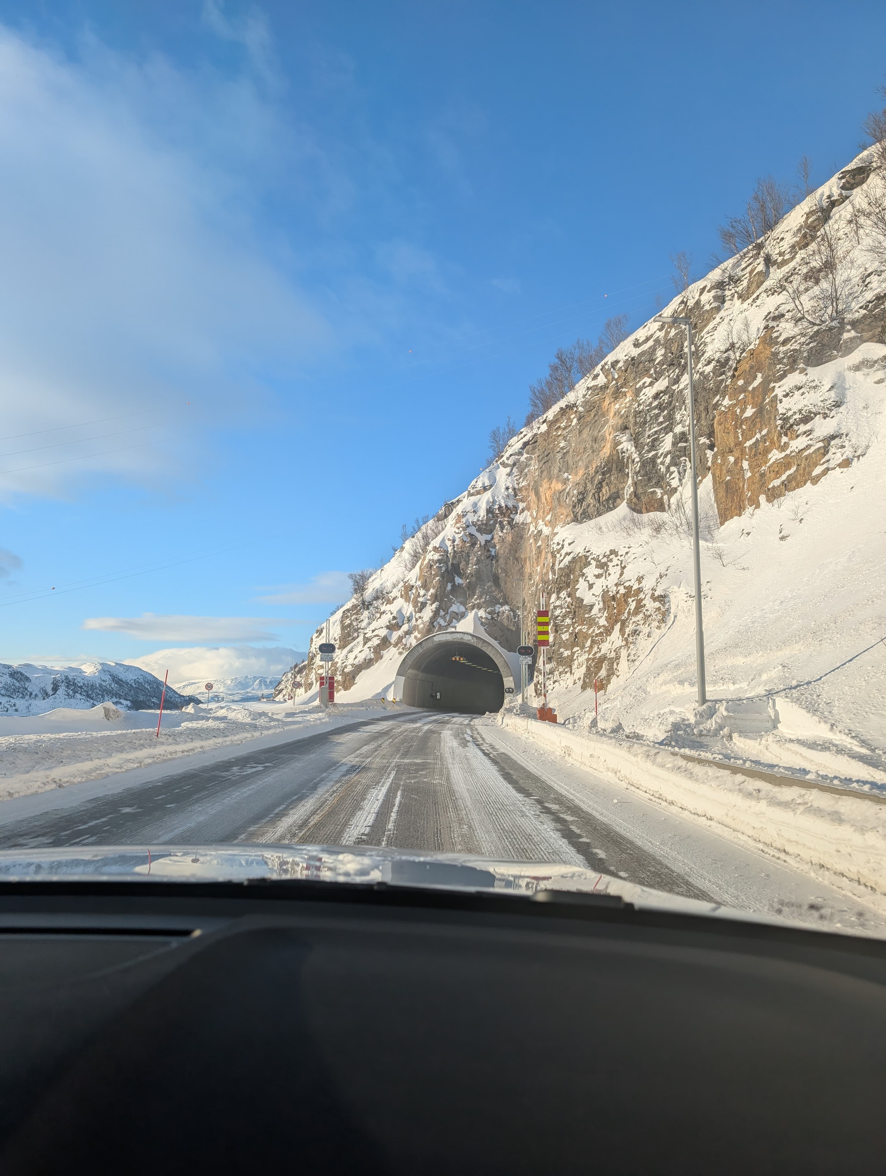 Eine verschneite Straße führt unter einem strahlend blauen Winterhimmel in einen Tunnel, eingebettet in eine steile Felslandschaft. Die Aufnahme vermittelt das Gefühl einer abenteuerlichen Autofahrt durch die norwegische Wildnis.