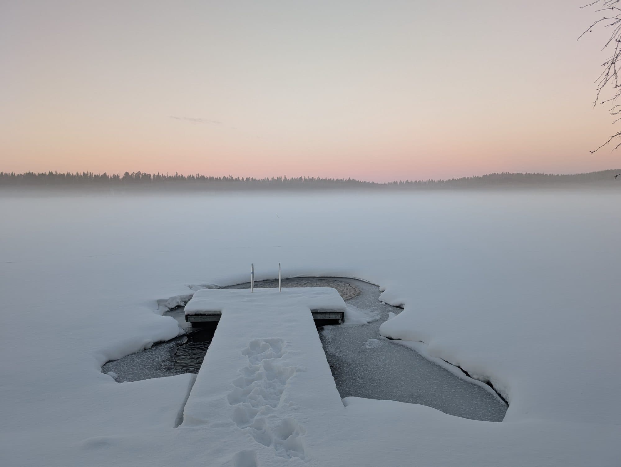 Ein schneebedeckter Holzsteg mit Metallleiter führt zu einer offenen Eisschwimmstelle im zugefrorenen See, umgeben von zartem Abendrosa am Horizont.