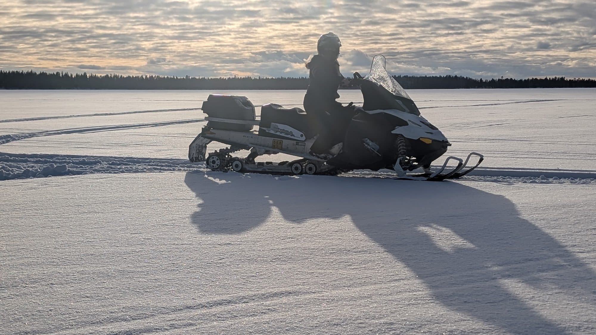 Eine Fahrerin durchquert im Gegenlicht mit dem Schneemobil einen weiten zugefrorenen See bei Kuusamo, im Hintergrund säumt dunkler Nadelwald den Horizont.