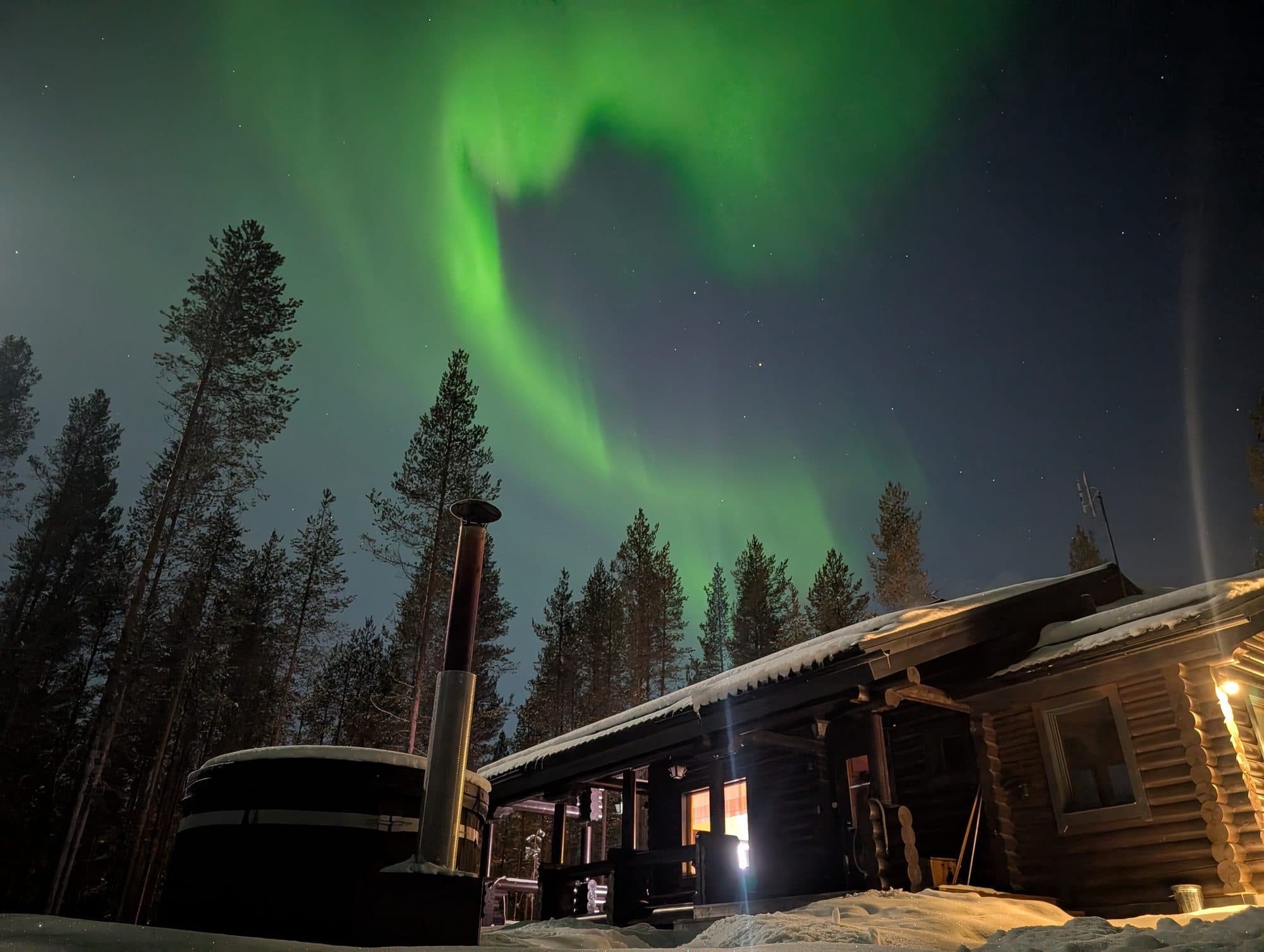 Über einer Blockhütte mit rauchendem Kamin und einem hölzernen Outdoor-Whirlpool im Schnee entfaltet sich ein leuchtend grünes Polarlicht am Nachthimmel von Kuusamo.
