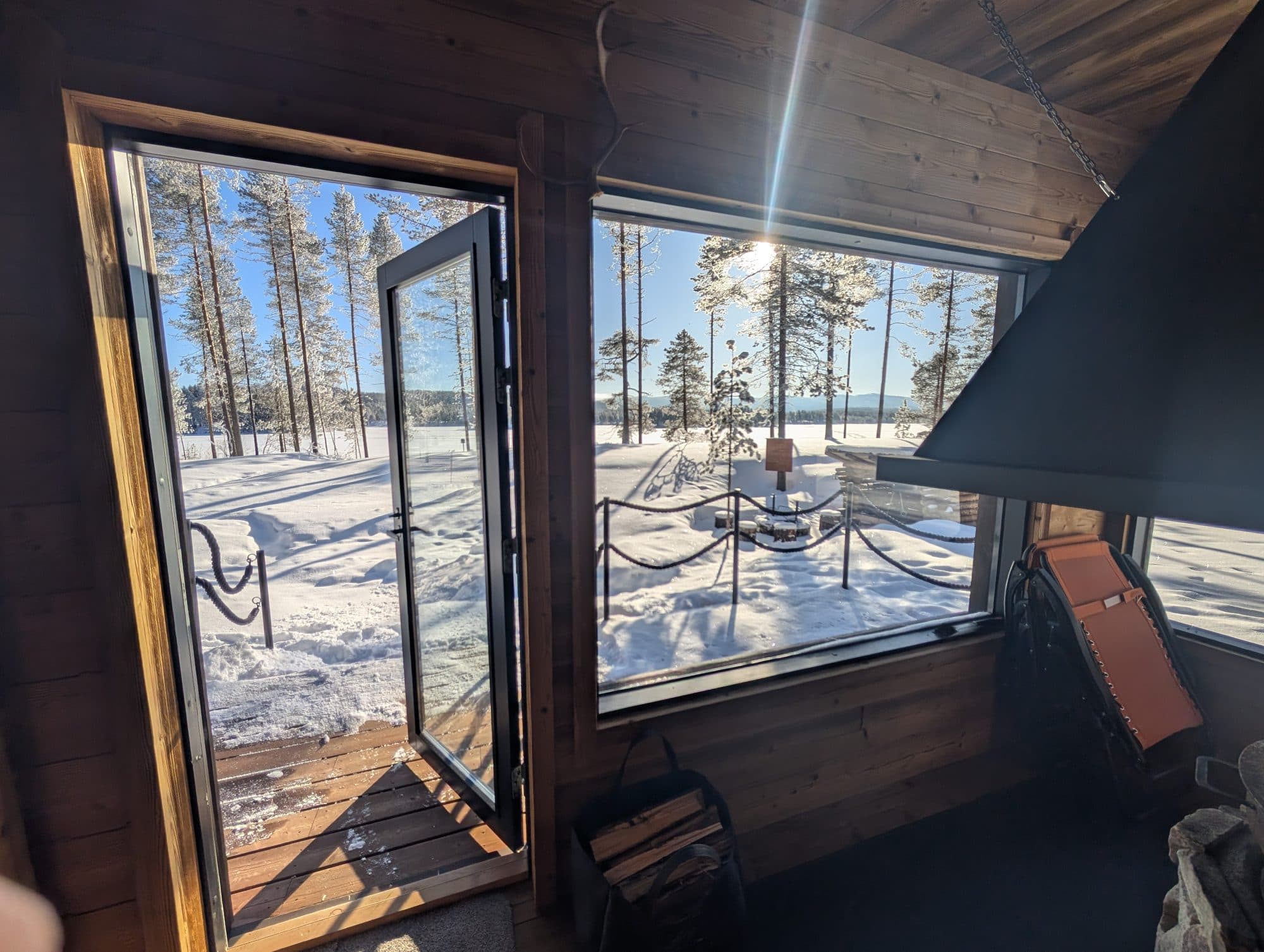 Blick aus der offenen Tür einer Feuerstellenhütte auf einen zugefrorenen See, umgeben von raureifbedeckten Kiefern im hellen Wintersonnenlicht.