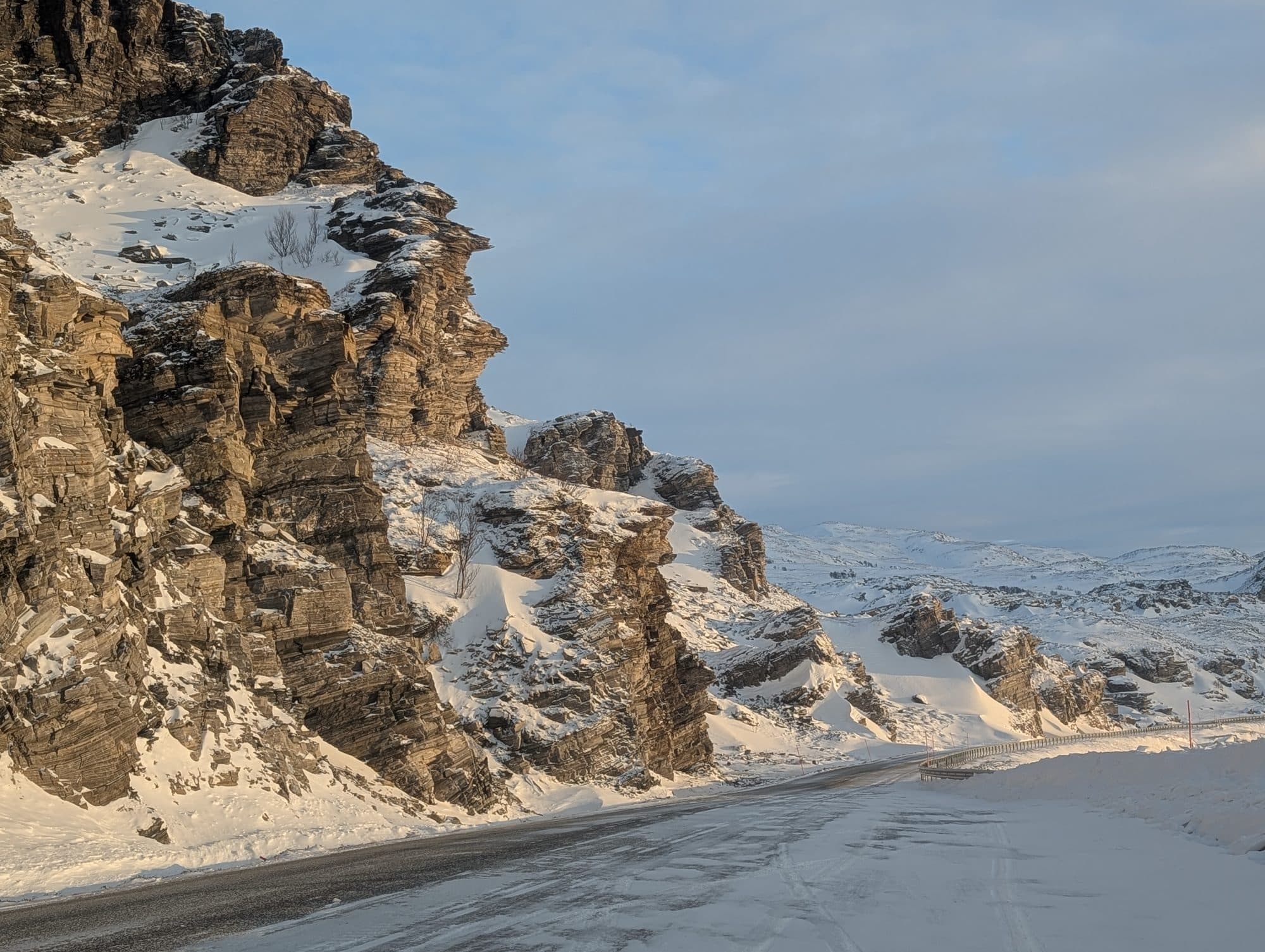 Eine schneebedeckte Straße schlängelt sich an steilen, sonnenbeschienenen Felswänden entlang in einer ruhigen Winterlandschaft.