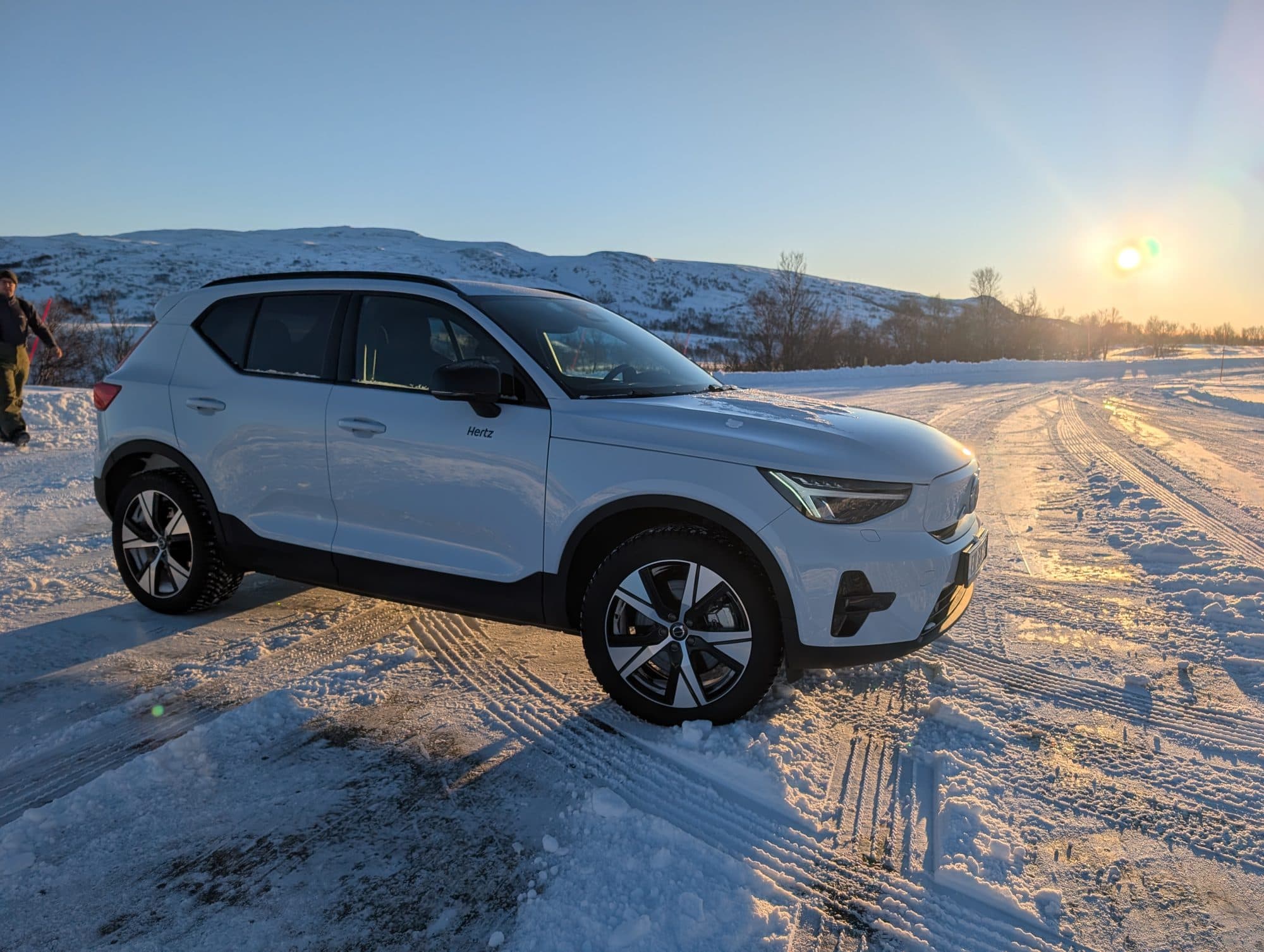 Ein weißer Volvo XC40 steht auf einer verschneiten Straße vor schneebedeckten Hügeln bei tiefstehender Wintersonne.