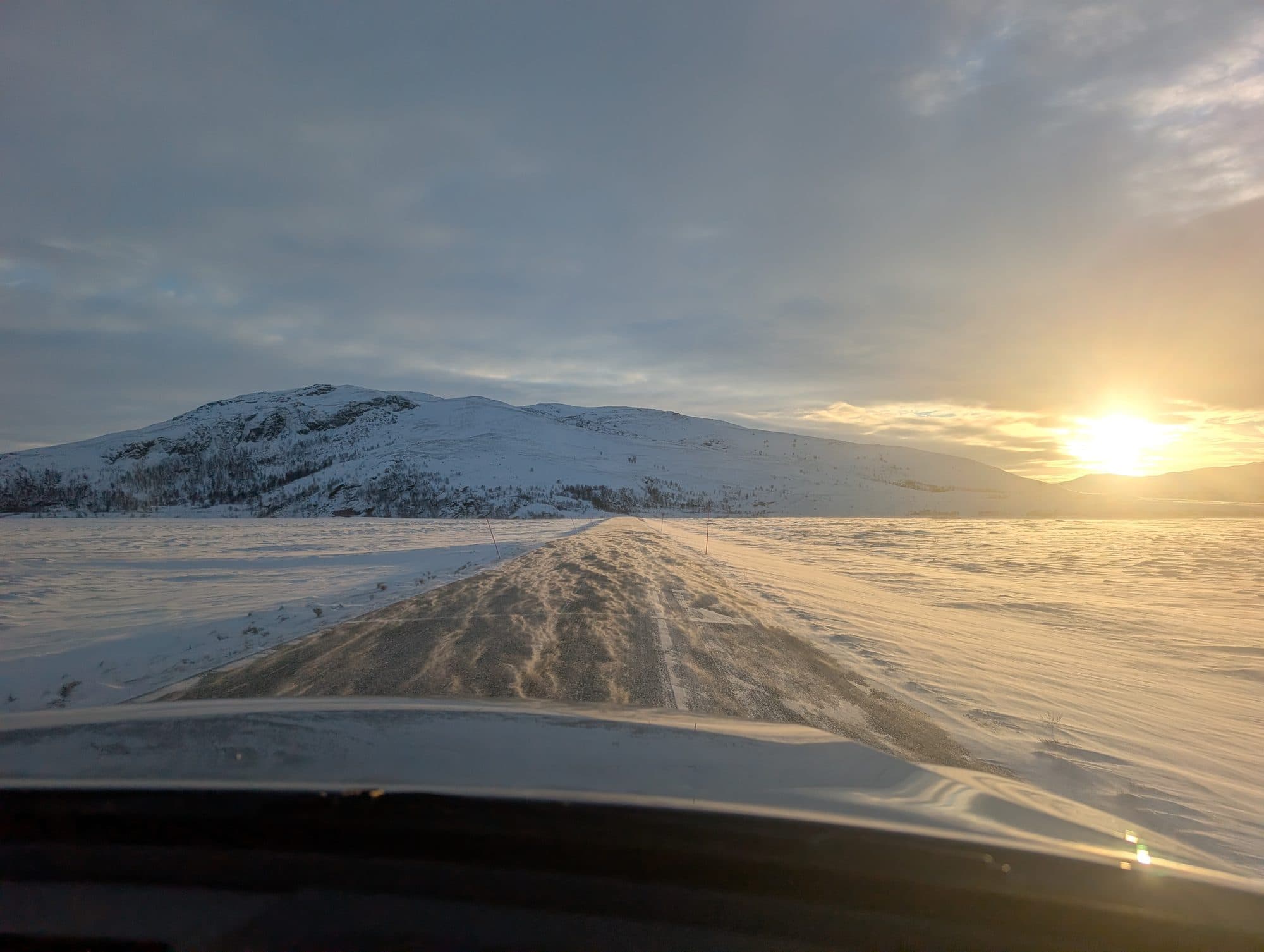 Blick aus dem Auto auf eine verschneite Straße, die sich durch eine weite Landschaft zu den Bergen zieht.