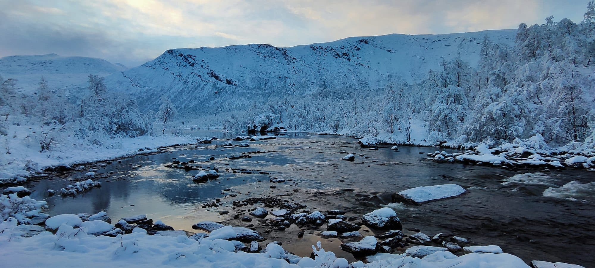 Panoramablick auf einen winterlichen Fluss mit schneebedeckten Felsen, umrahmt von verschneiten Bergen.