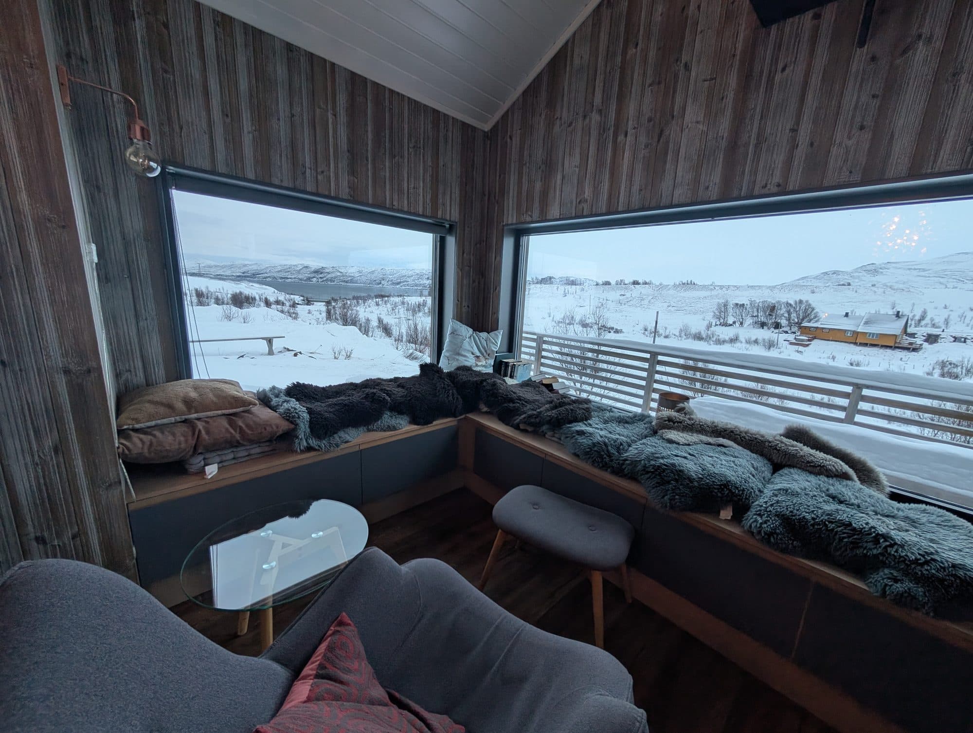 Modernes Ferienhaus-Interieur mit Panoramafenstern, Fellen auf der Sitzbank und Fjordblick im Winter.