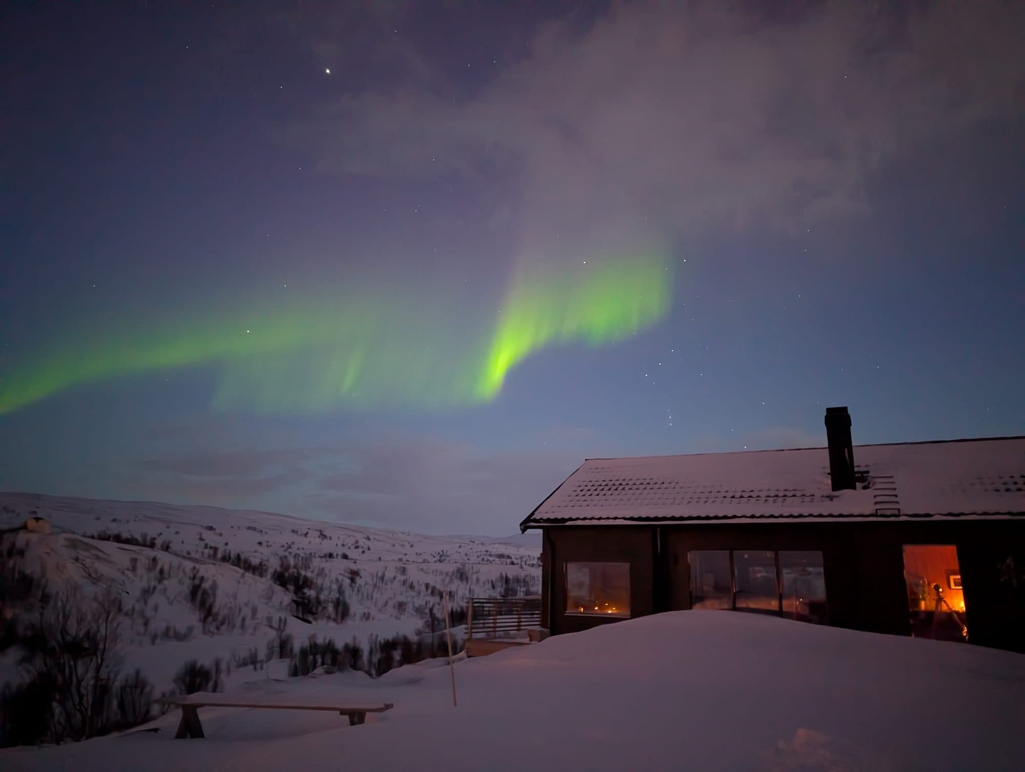 Intensiv grüne Nordlichter bilden dramatische Vorhänge über dem beleuchteten Ferienhaus.