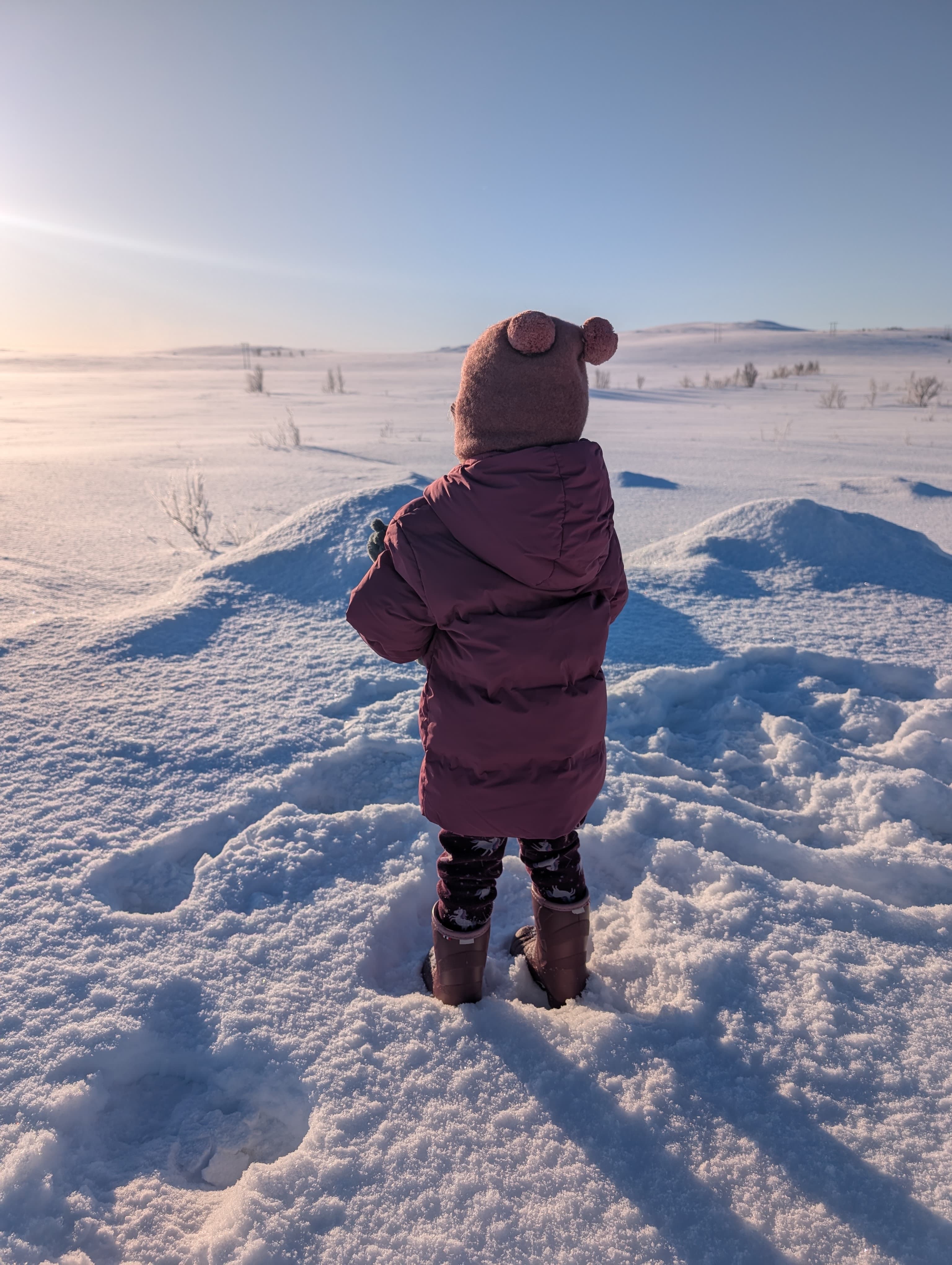 Ein Kind in winterlicher Kleidung steht mit dem Rücken zur Kamera und blickt über eine weite, schneebedeckte Landschaft bei tiefstehender Sonne.