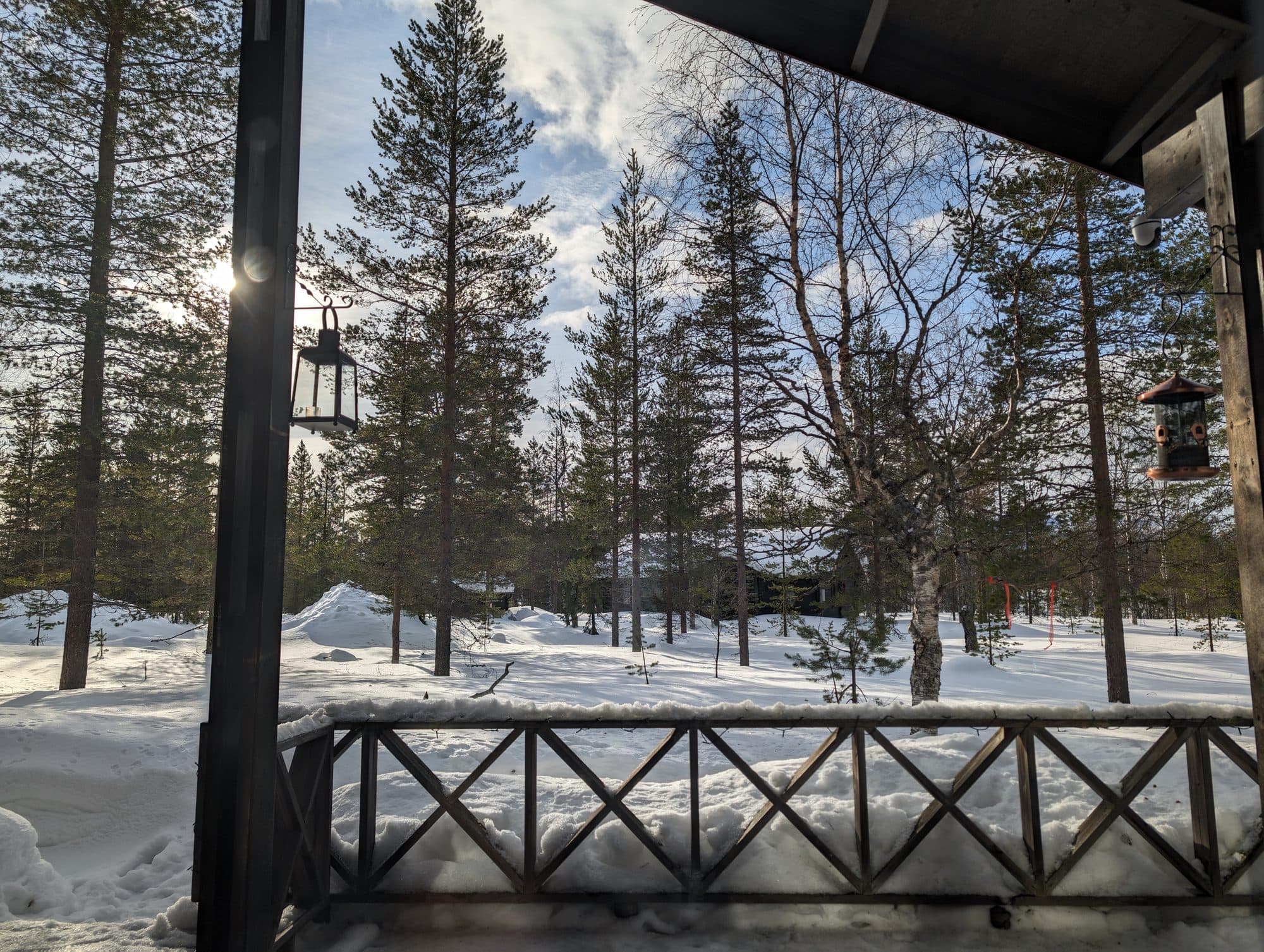 Blick von der Veranda einer Ferienhütte über ein verschneites Waldgrundstück mit Holzgeländer, Laterne und Vogelfutterhaus.