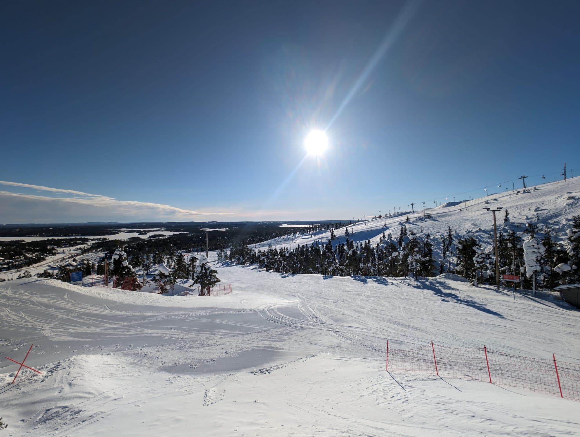 Ein sonniger Wintertag in Kuusamo, Finnland, zeigt eine weite, schneebedeckte Skipiste mit Skiliften und einer malerischen Winterlandschaft aus Wäldern und zugefrorenen Seen im Hintergrund.