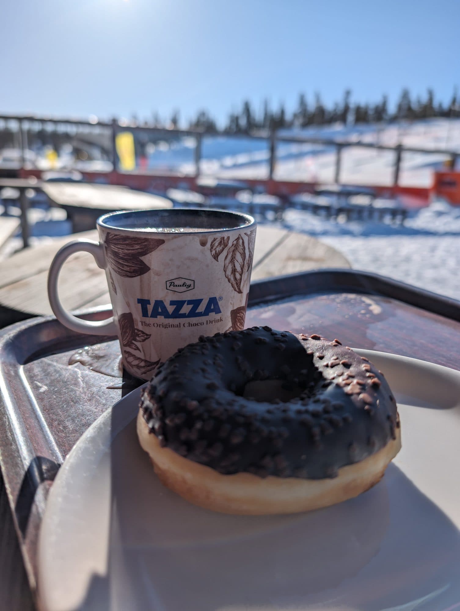 Schokoladen-Donut und eine Tasse heiße Schokolade auf einem Tablett, im unscharfen Hintergrund die sonnige Skipiste von Ruka – eine typische Après-Ski-Pause.