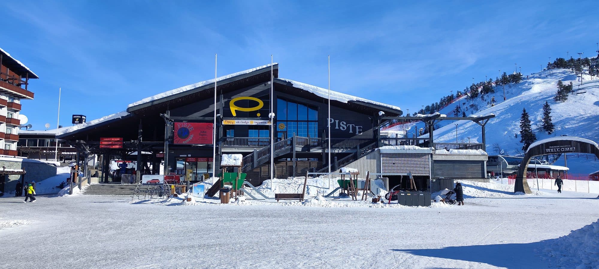 Das Piste Ski Lodge am Fuß der Pisten von Ruka, mit Blick auf die schneebedeckten Hänge und den Skilift im Hintergrund.