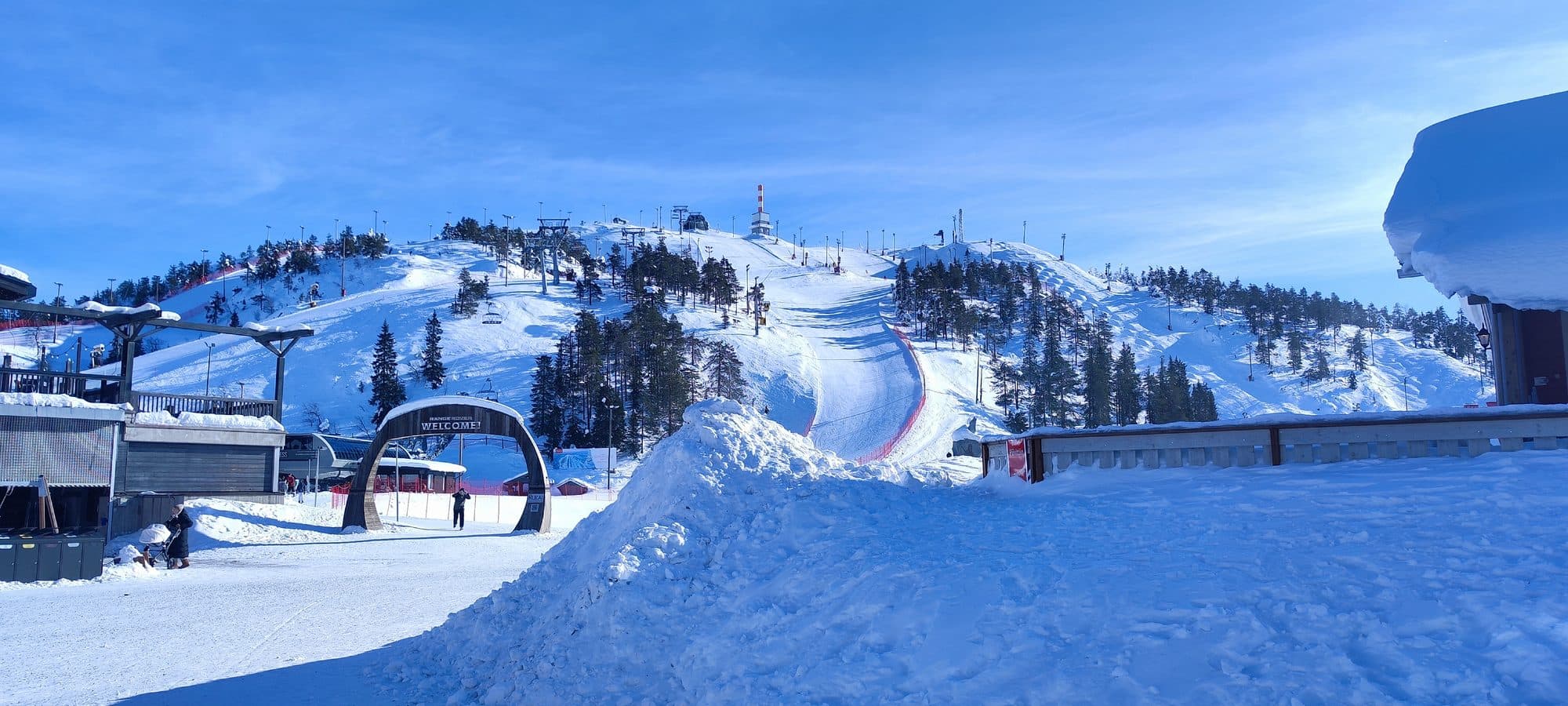 Panoramablick auf die Skipisten von Ruka mit hölzernem Welcome-Torbogen im Vordergrund und Skiliften, die den Berg hinaufführen.