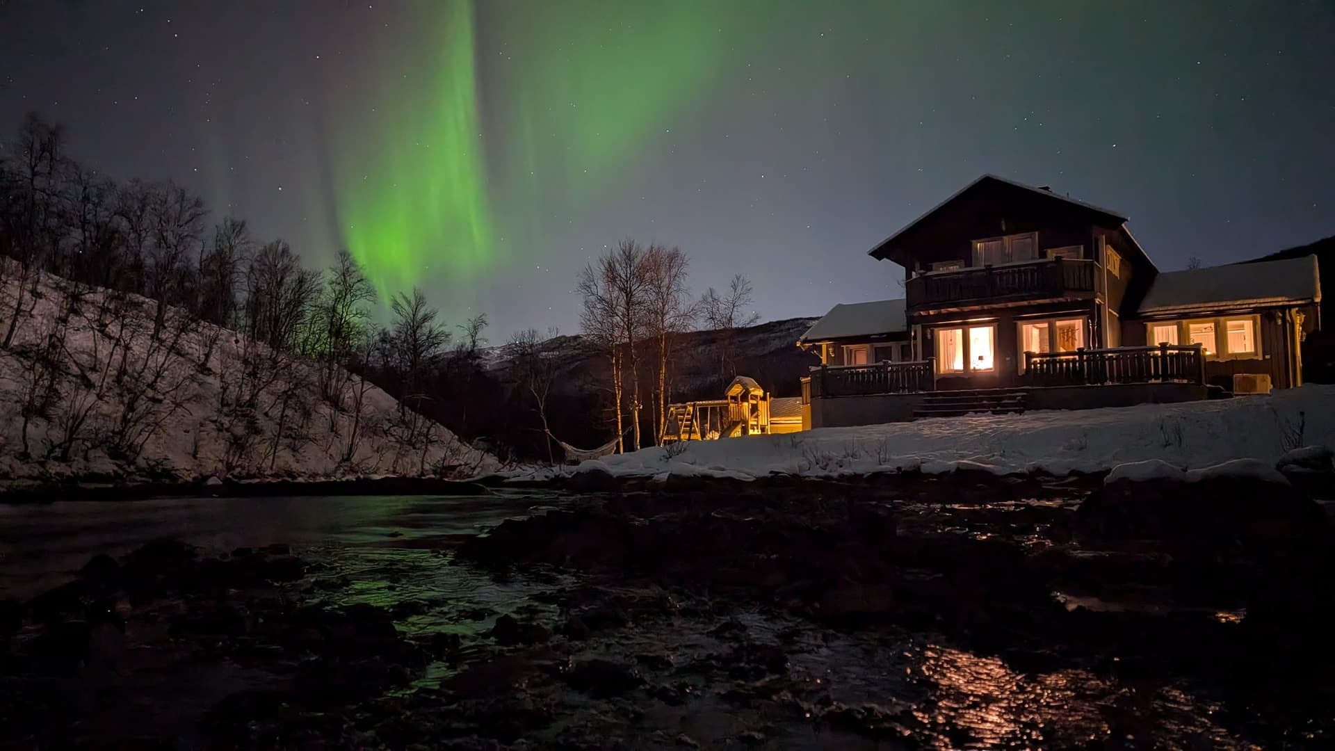 Intensives grünes Polarlicht wallt über einem gemütlich beleuchteten Holzchalet mit Veranda, im Vordergrund ein dunkler Fluss und eine schneebedeckte Böschung.