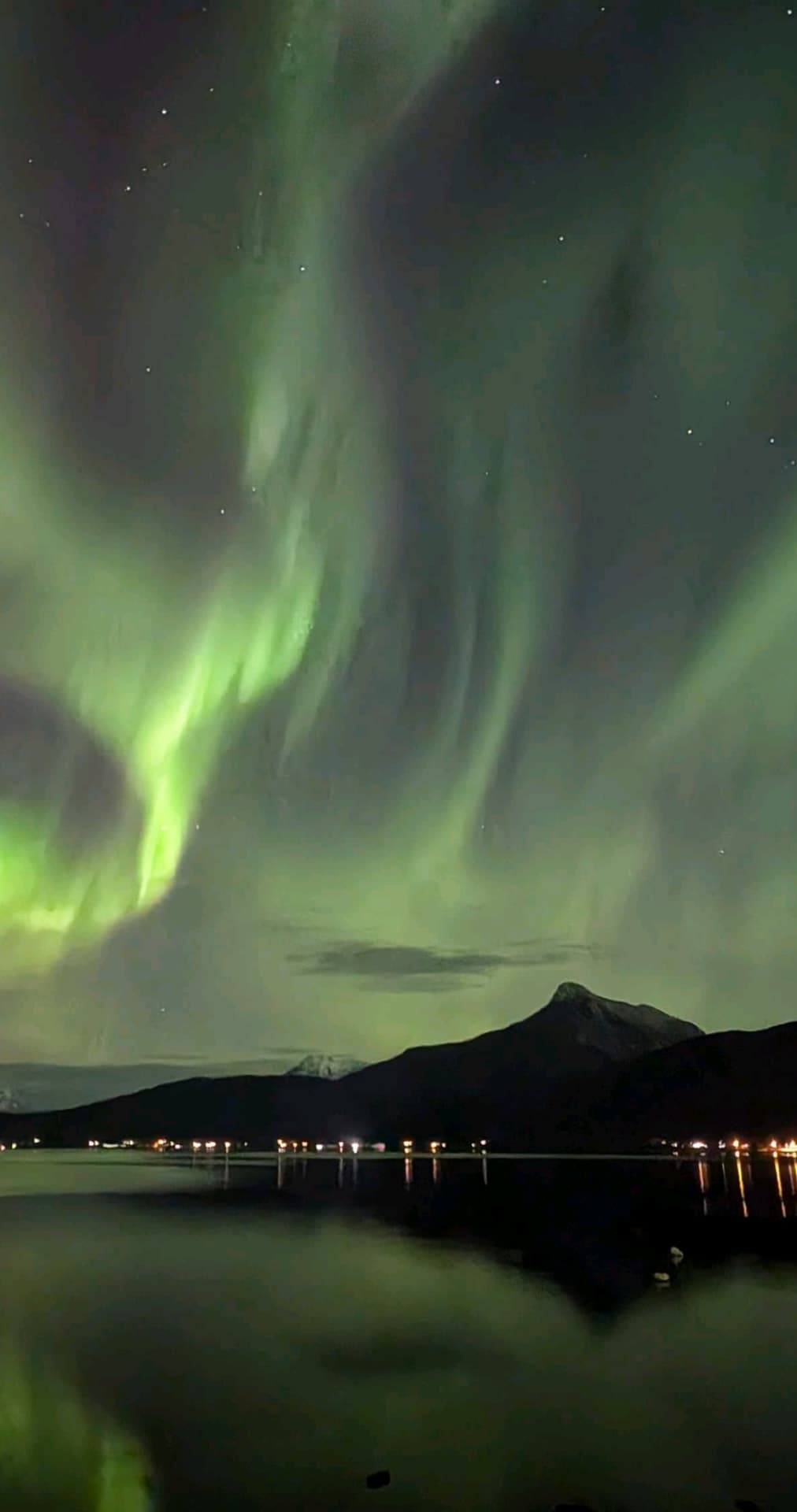 Intensives grünes Polarlicht tanzt über einem nächtlichen Fjord bei Ballangen und spiegelt sich im ruhigen Wasser, während am Ufer vereinzelt Lichter eines kleinen Ortes leuchten.