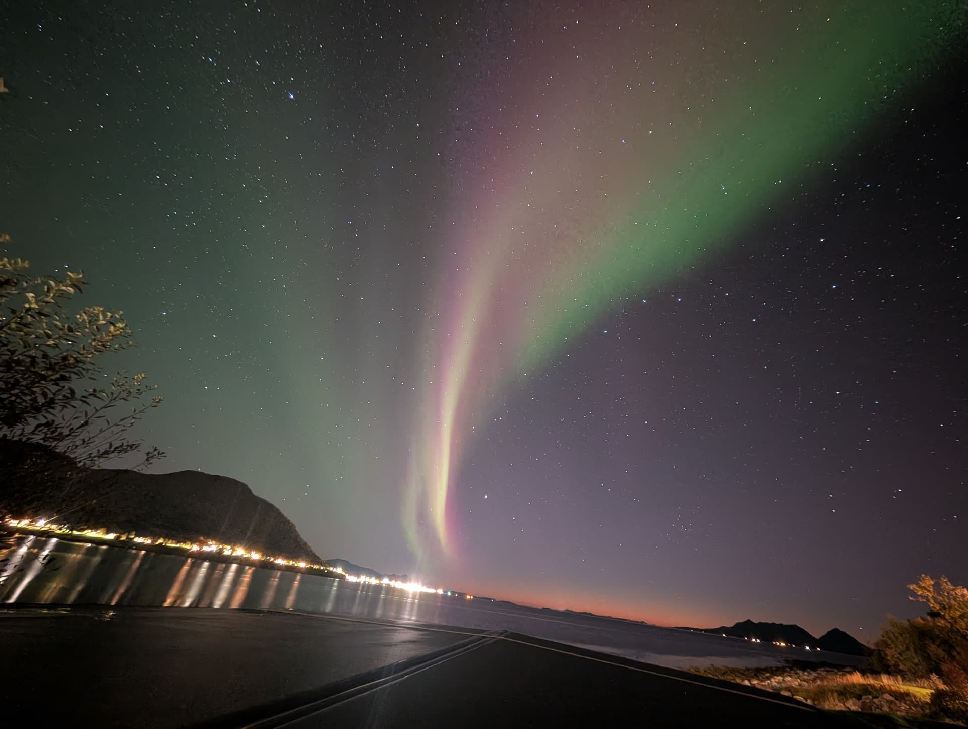 Zweite Aufnahme desselben spektakulären grün-violetten Polarlichts über der Küstenstraße am Fjord bei Andørja mit gespiegelten Uferlichtern und rötlichem Dämmerungsstreifen am Horizont.