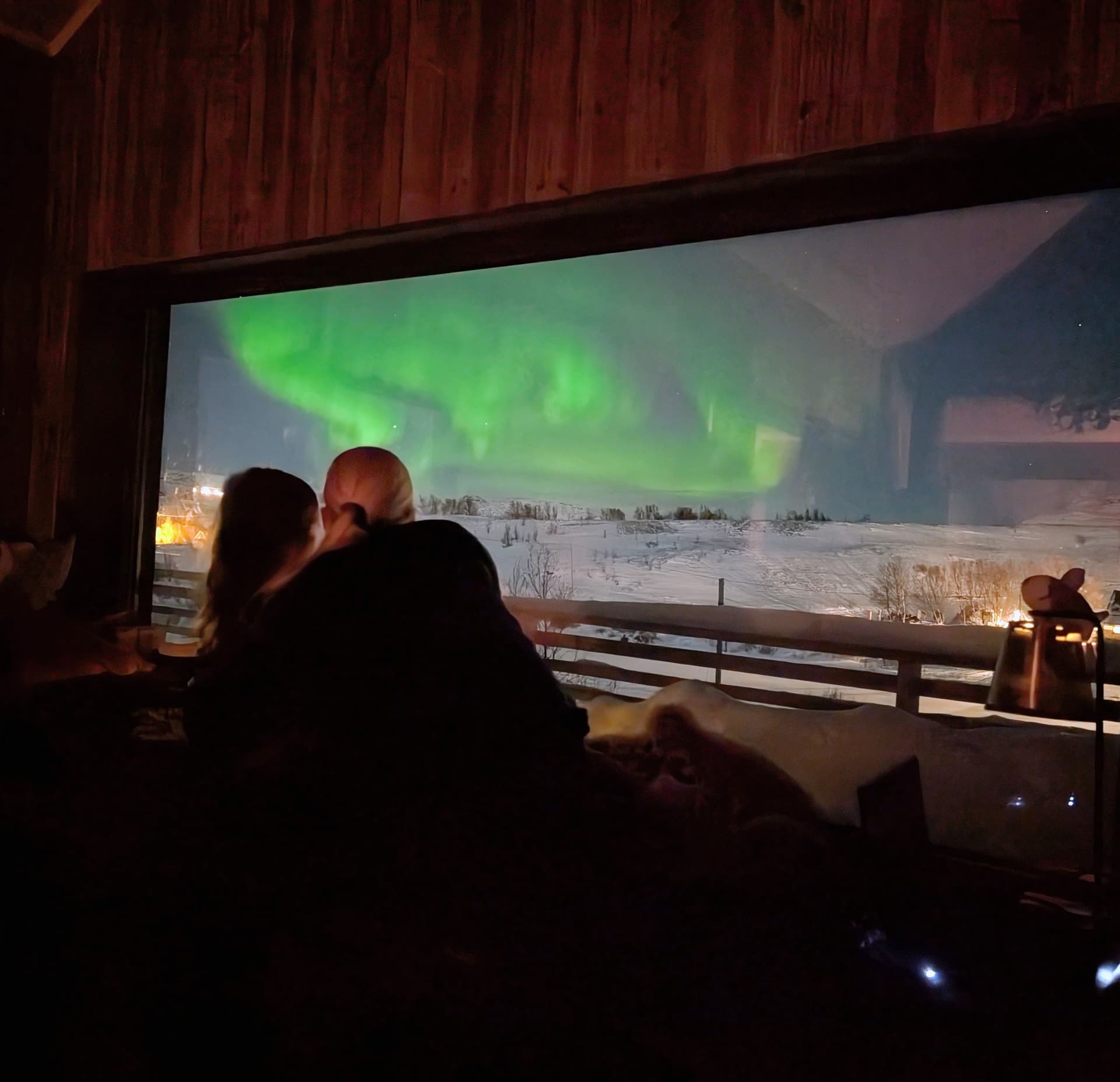 Zwei Personen blicken von einem dunklen, holzgetäfelten Raum durch ein großes Fenster auf leuchtende grüne Polarlichter über einer verschneiten Landschaft.