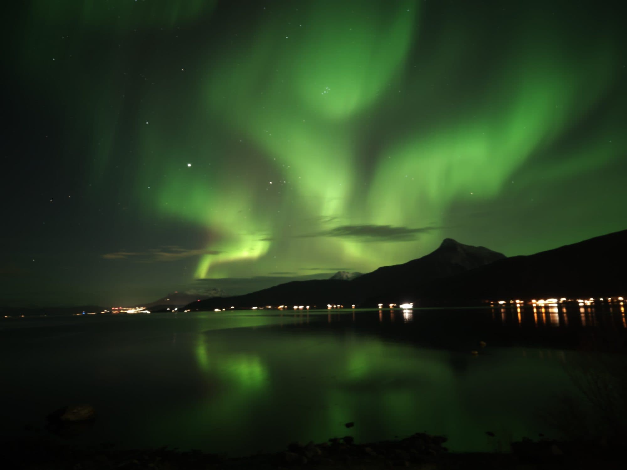 Ein leuchtend grünes Polarlicht mit mehreren strahlenförmigen Bändern zieht über einen Fjord bei Ballangen und spiegelt sich im spiegelglatten Wasser, während am Ufer die Lichter eines kleinen Küstenortes vor dunkler Bergsilhouette funkeln.