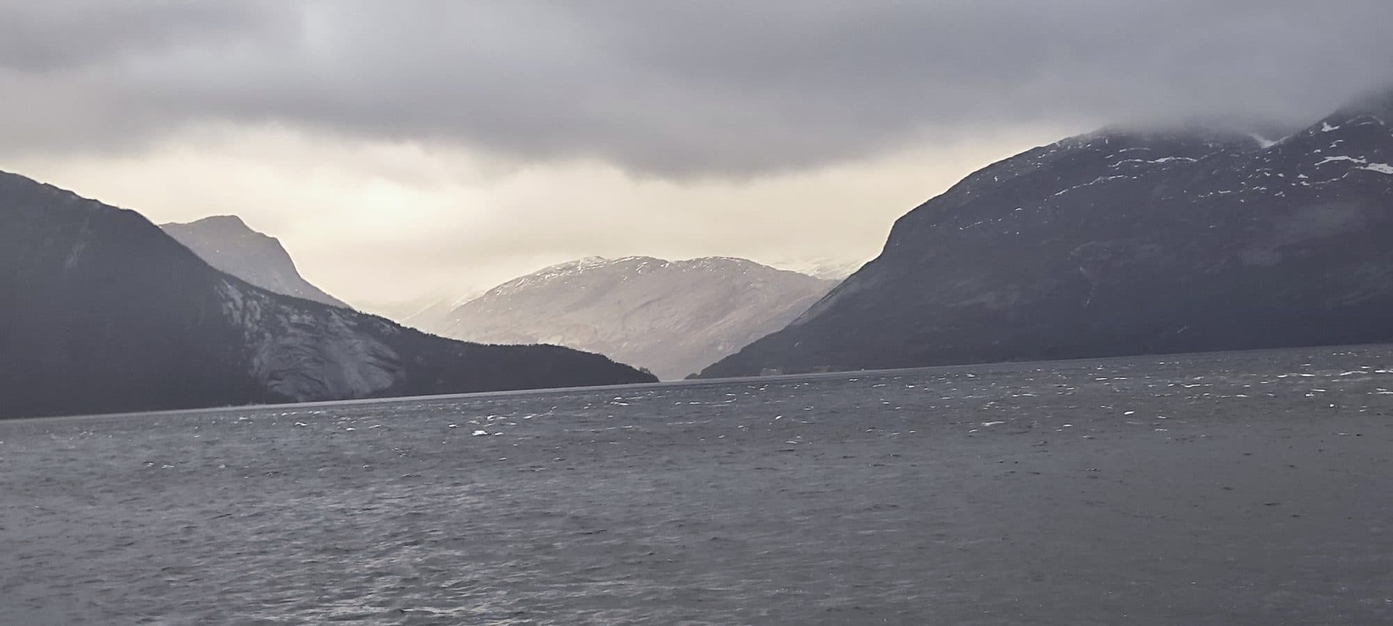 Blick über einen Fjord bei Ballangen, während die Wintersonne durch aufreißende Wolken einen fernen Berghang beleuchtet.
