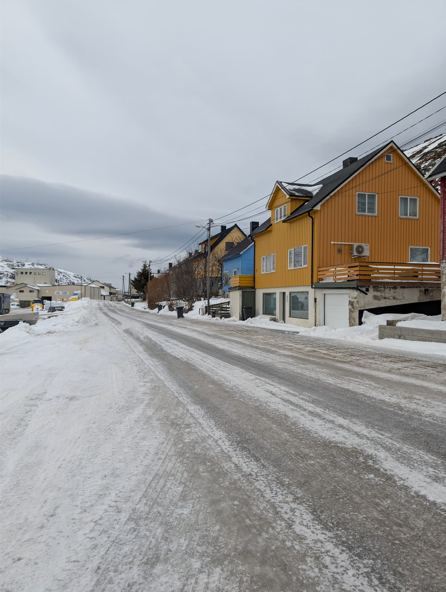 Eine verschneite Straße führt durch ein farbenfrohes norwegisches Fischerdorf unter einem grauen Winterhimmel. Die Szene vermittelt eine ruhige, authentische Winteratmosphäre im Norden.