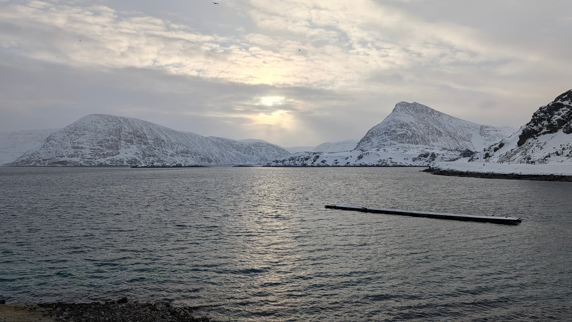 Eine ruhige winterliche Szene in Nordnorwegen mit schneebedeckten Bergen und einem Holzsteg, der ins dunkle Wasser ragt. Die Sonne bricht schwach durch die Wolken und erzeugt eine kühle, friedliche Atmosphäre.
