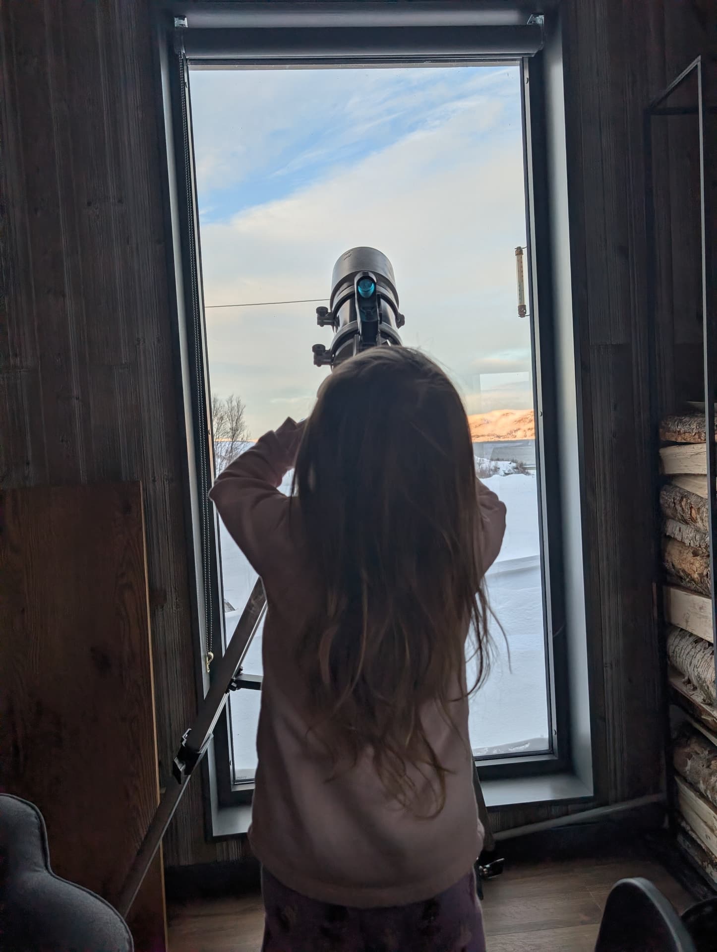 Kind schaut durch Teleskop am Fenster, Schnee und Fass-Sauna draußen sichtbar.