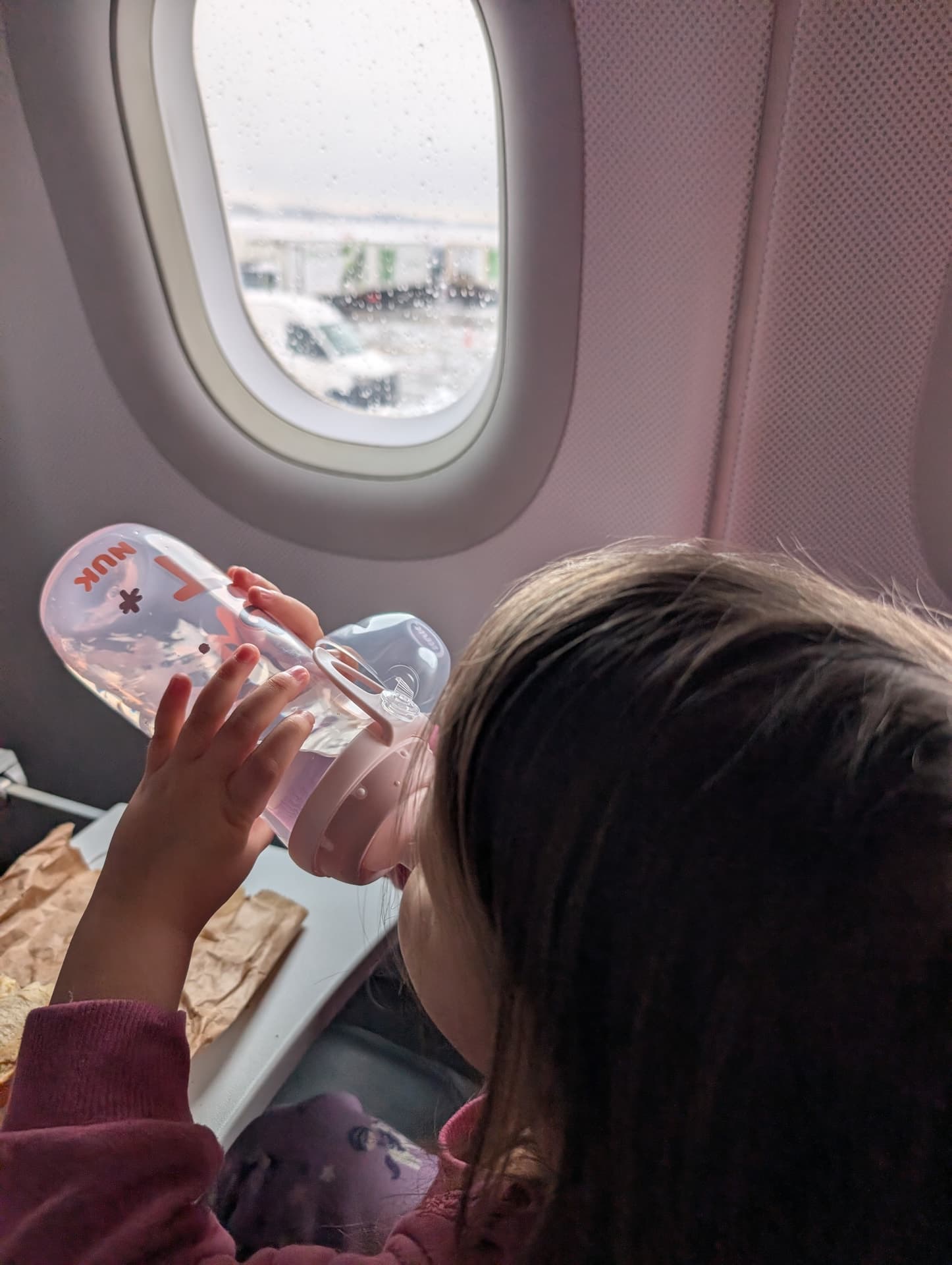 Ein Kind sitzt im Flugzeug und schaut neugierig aus dem Fenster, während es aus einer Trinkflasche trinkt. Die Stimmung ist ruhig und erfüllt von Vorfreude auf die bevorstehende Reise.
