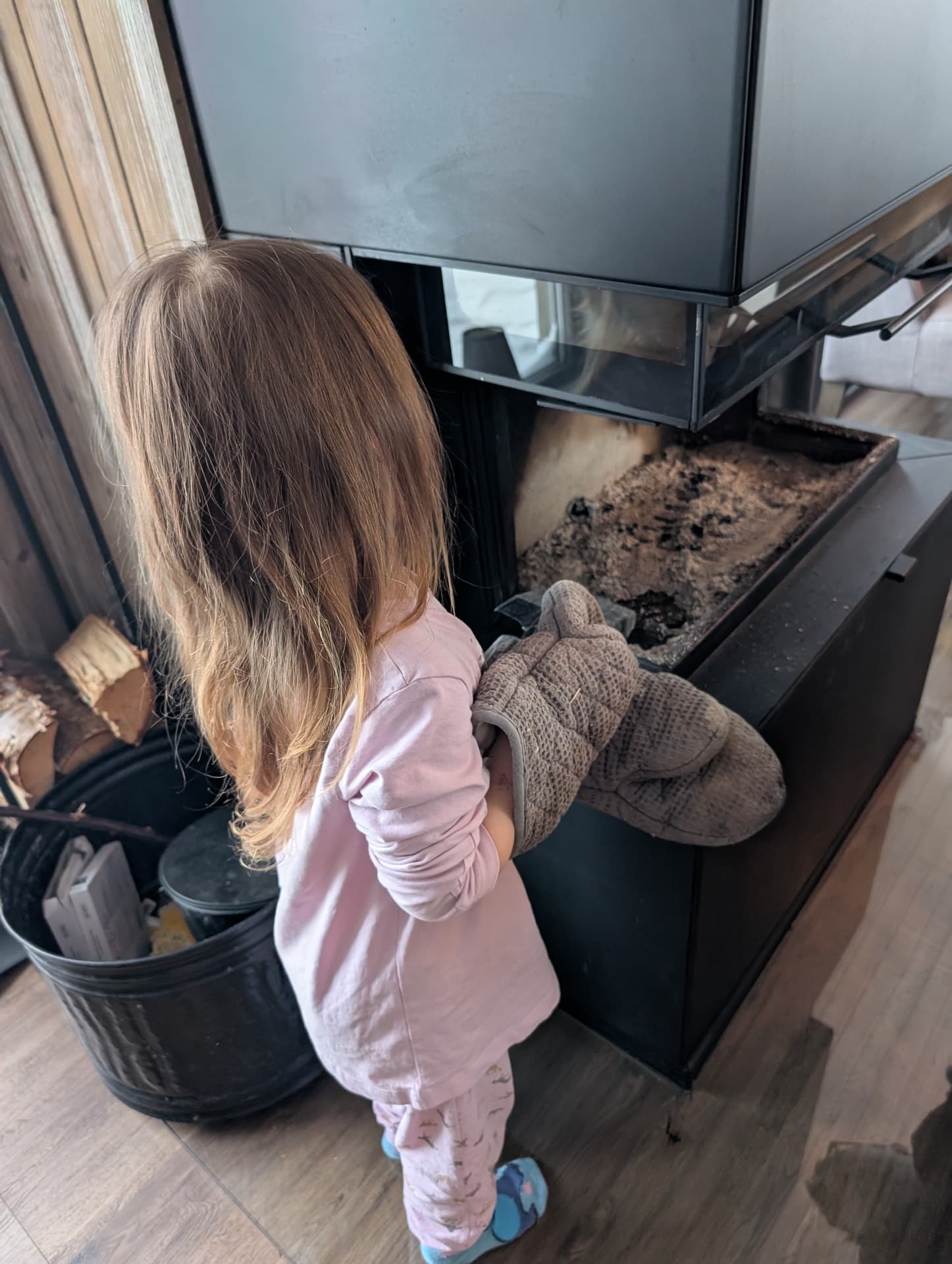 Ein kleines Mädchen in rosa Kleidung steht vor einem schwarzen Kaminofen und hält einen dicken Ofenhandschuh bereit, um Holz nachzulegen. Die Szene strahlt eine warme, winterliche Häuslichkeit aus.