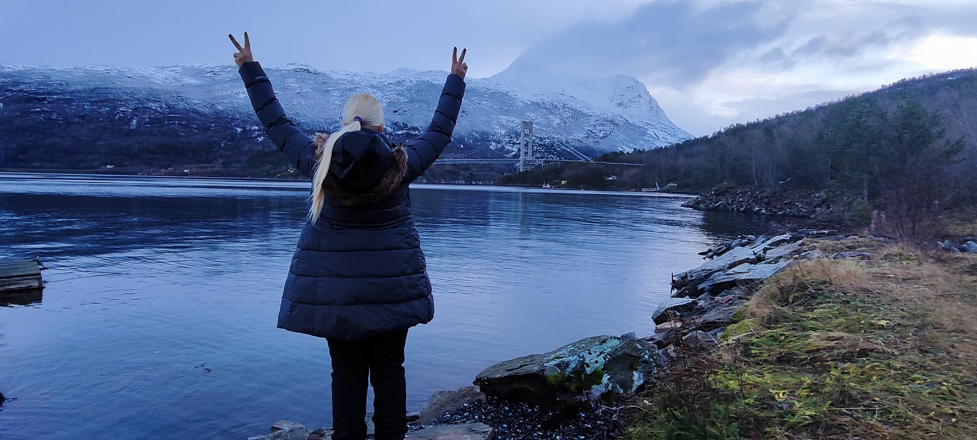 Eine Frau mit blondem Zopf reckt jubelnd beide Hände zum Peace-Zeichen in die Luft, vor ihr liegt der Fjord bei Narvik mit der Hålogalandsbrücke und schneebedeckten Bergen.