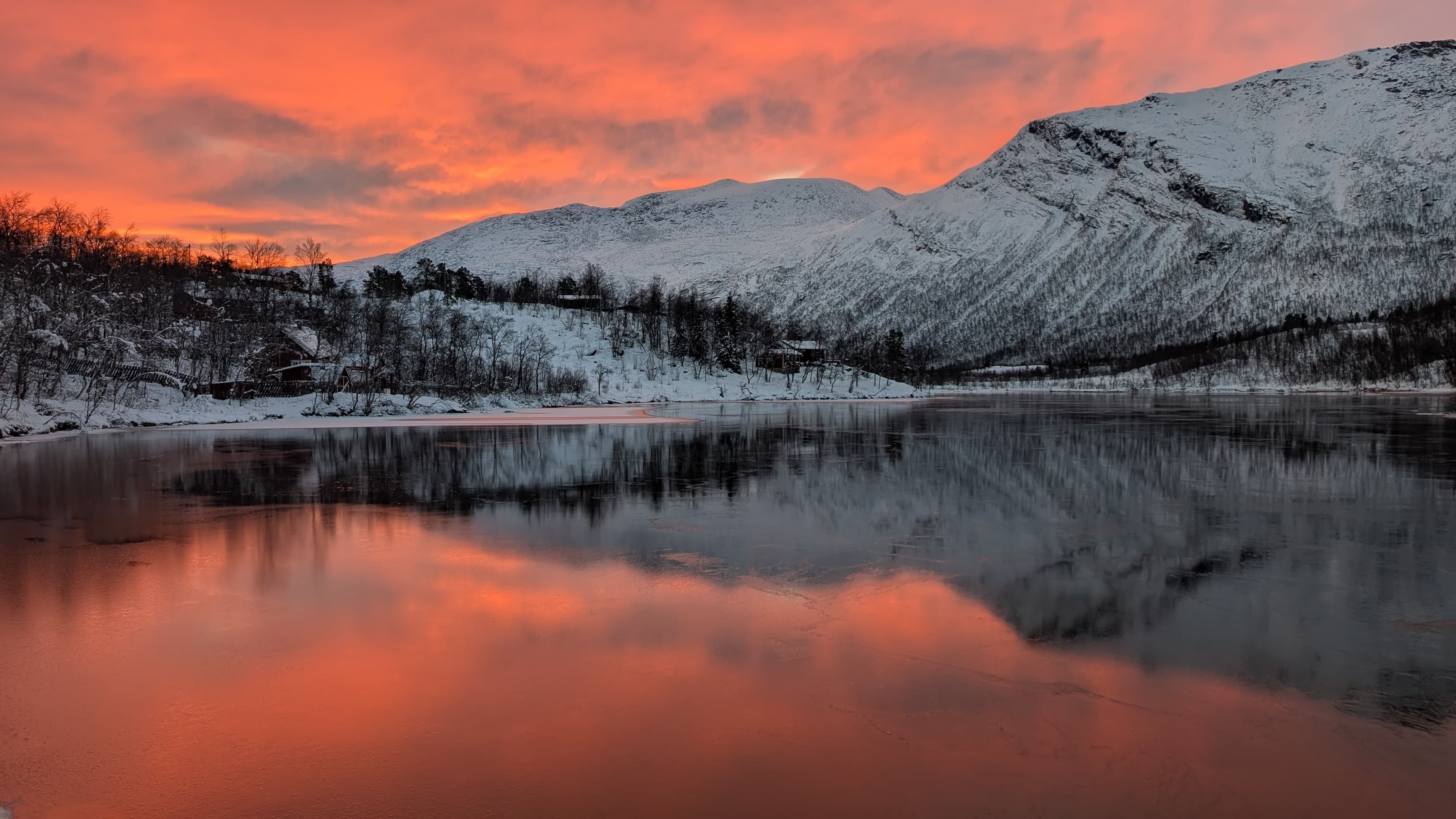 Panoramablick auf einen zugefrorenen See mit Eisschollen im Vordergrund, gerahmt von schneebedeckten Gipfeln unter glühend rotem Abendhimmel bei Narvik.
