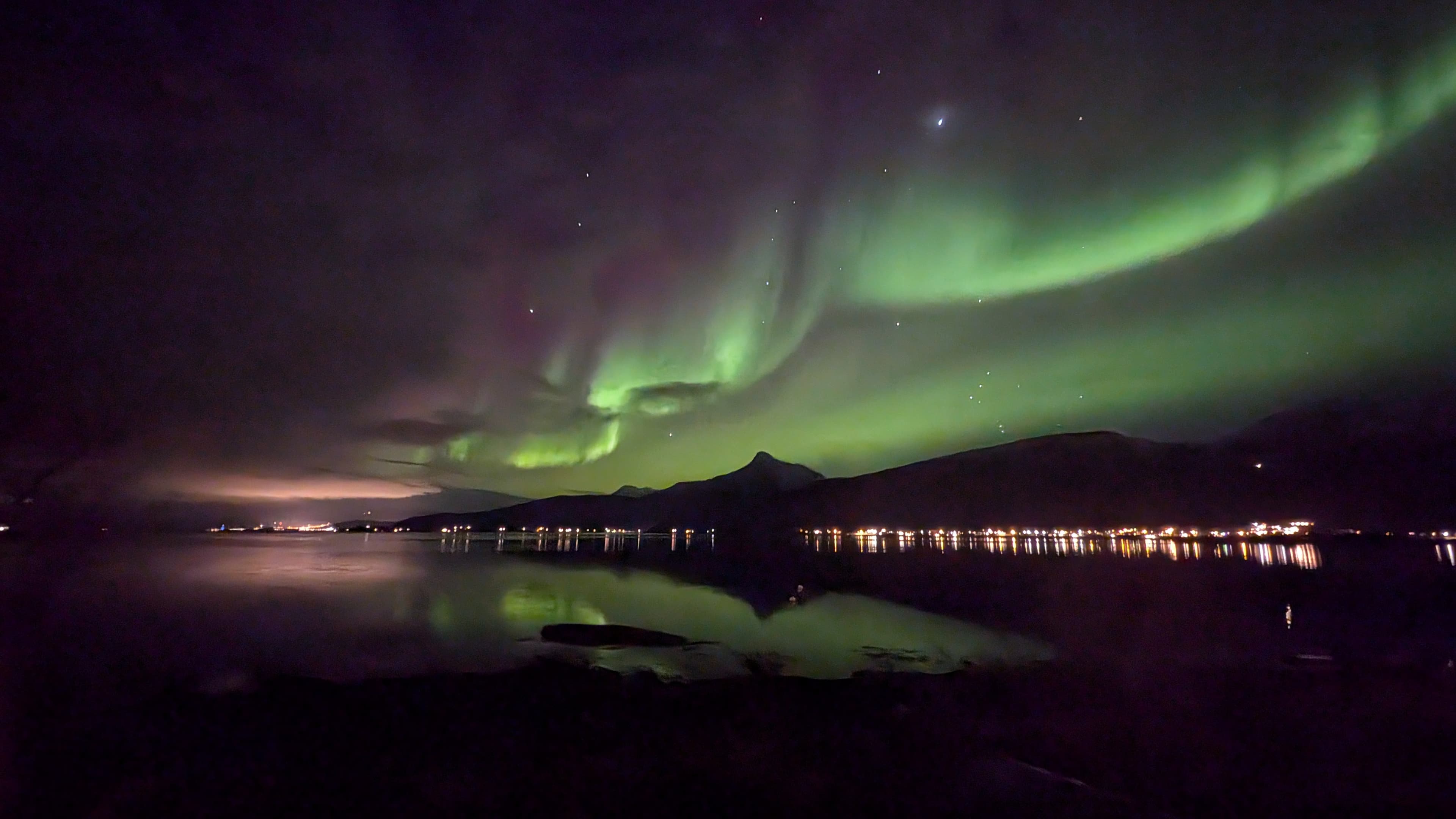 Ein leuchtend grünes Polarlicht mit violettem Schimmer schwingt in einer weiten Kurve über einen markanten Berggipfel am Ofotfjord, während sich die Lichter des Küstenorts im ruhigen Wasser spiegeln.