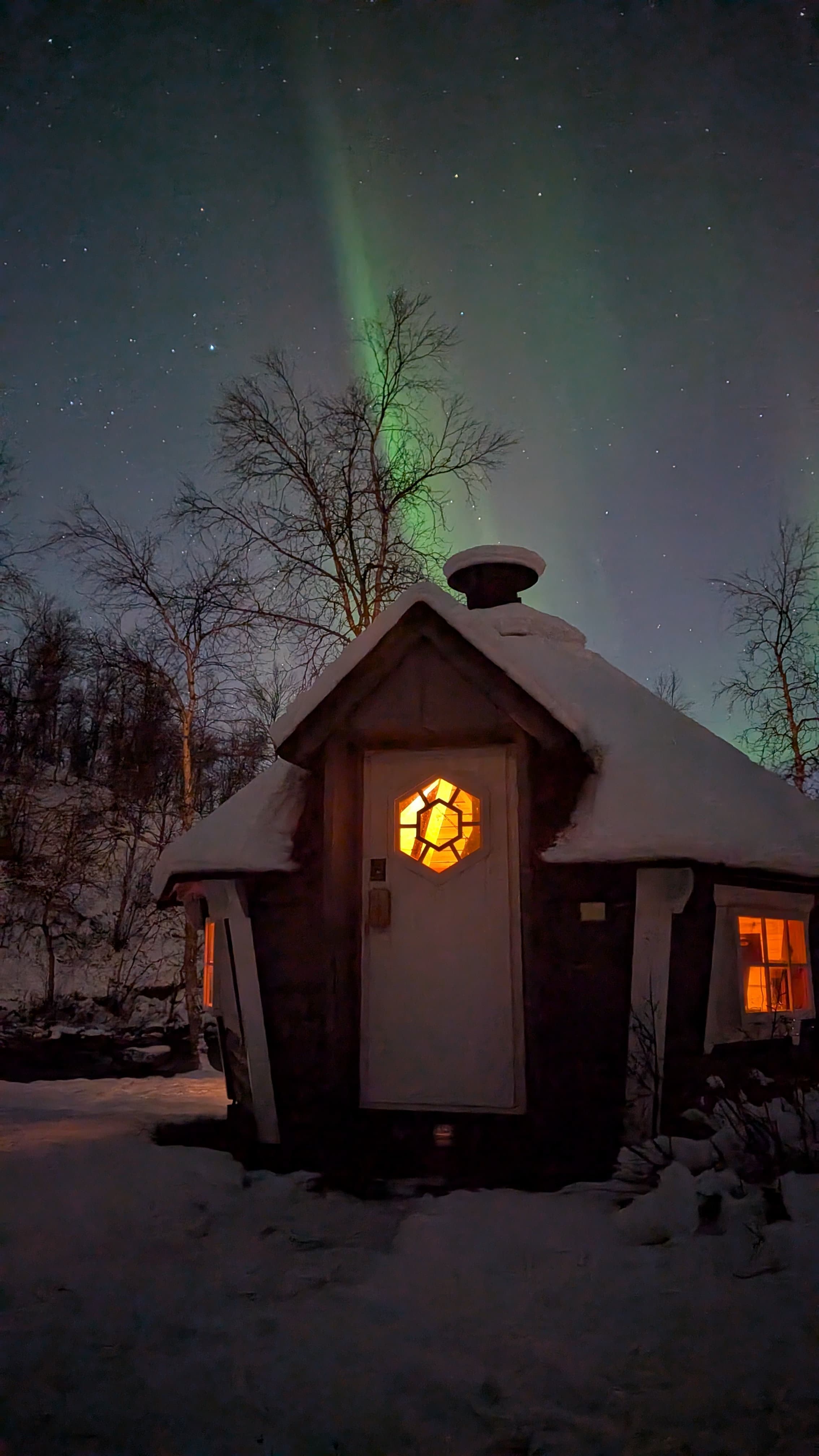 Ein schneebedecktes Holzhaus mit warm erleuchtetem Fenster steht unter einem intensiven grünen Polarlichtband am klaren Sternenhimmel bei Narvik.