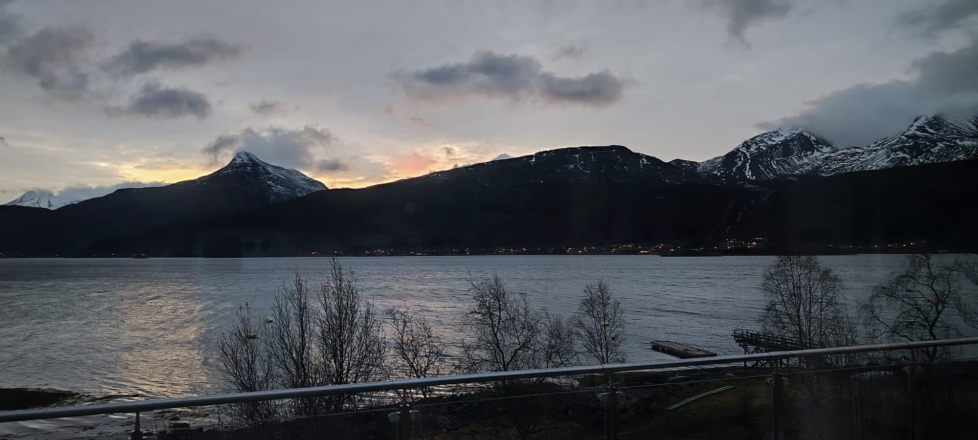 Weites Panorama des Fjords bei Ballangen in der Morgendämmerung mit schneebedeckten Bergketten und einem kleinen Bootssteg.