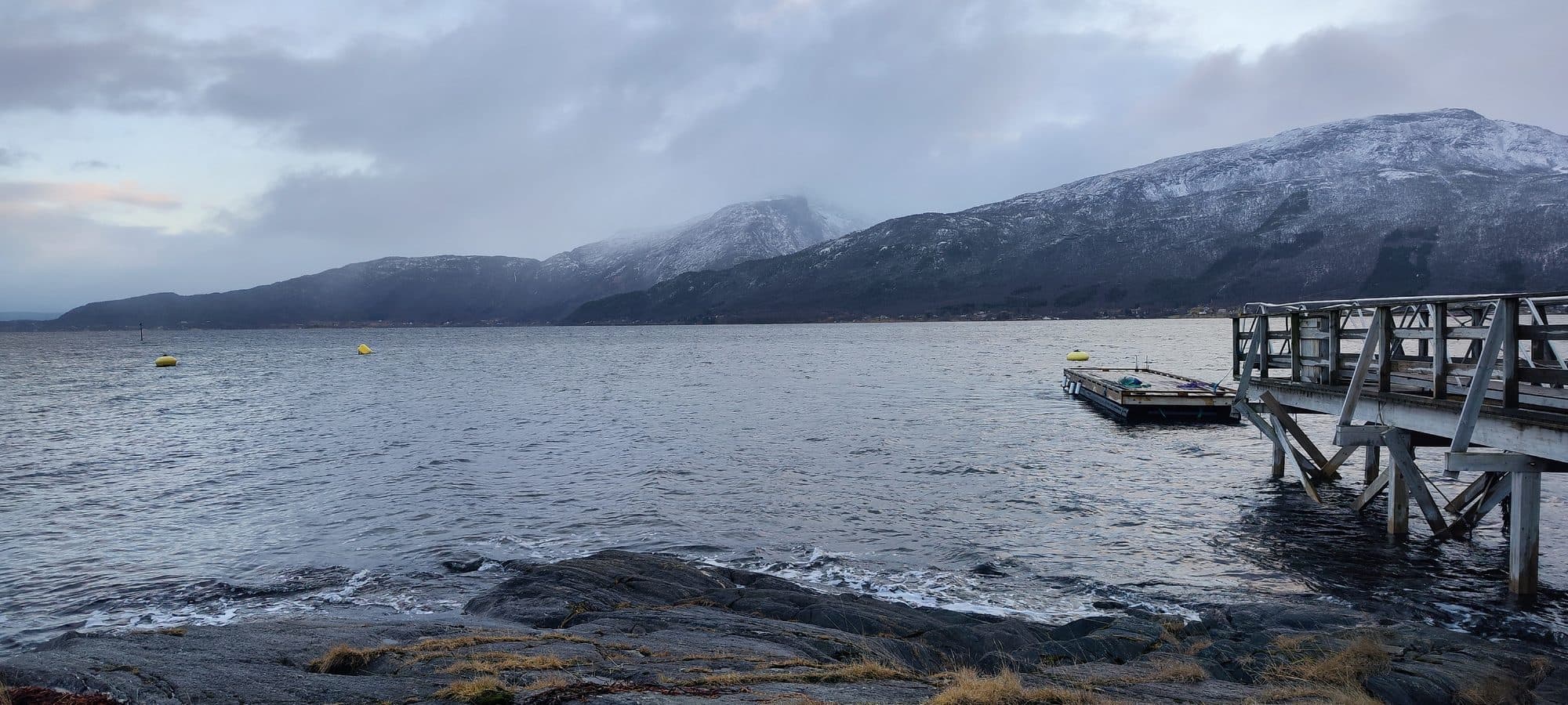 Blick über einen ruhigen Fjord bei Ballangen auf schneebedeckte Berge unter dichter Wolkendecke.