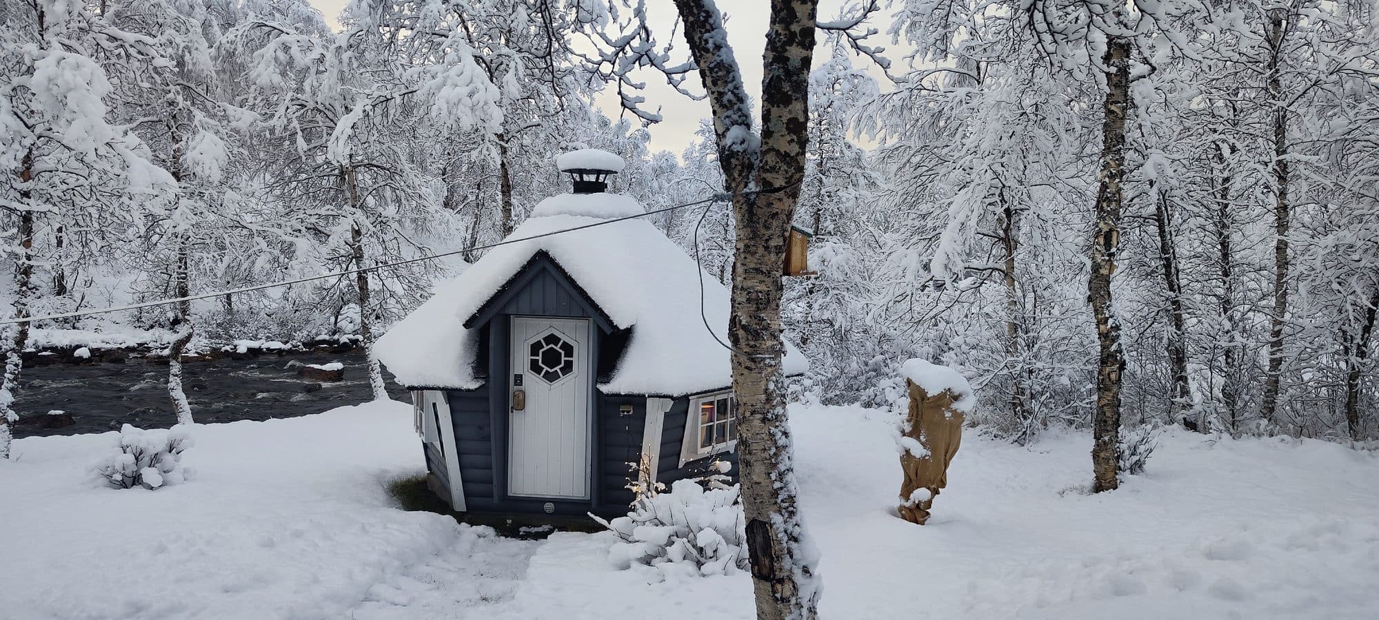 Eine kleine blaue Holzhütte mit schneebedecktem Dach und rundem Kaminaufsatz zwischen verschneiten Birken.