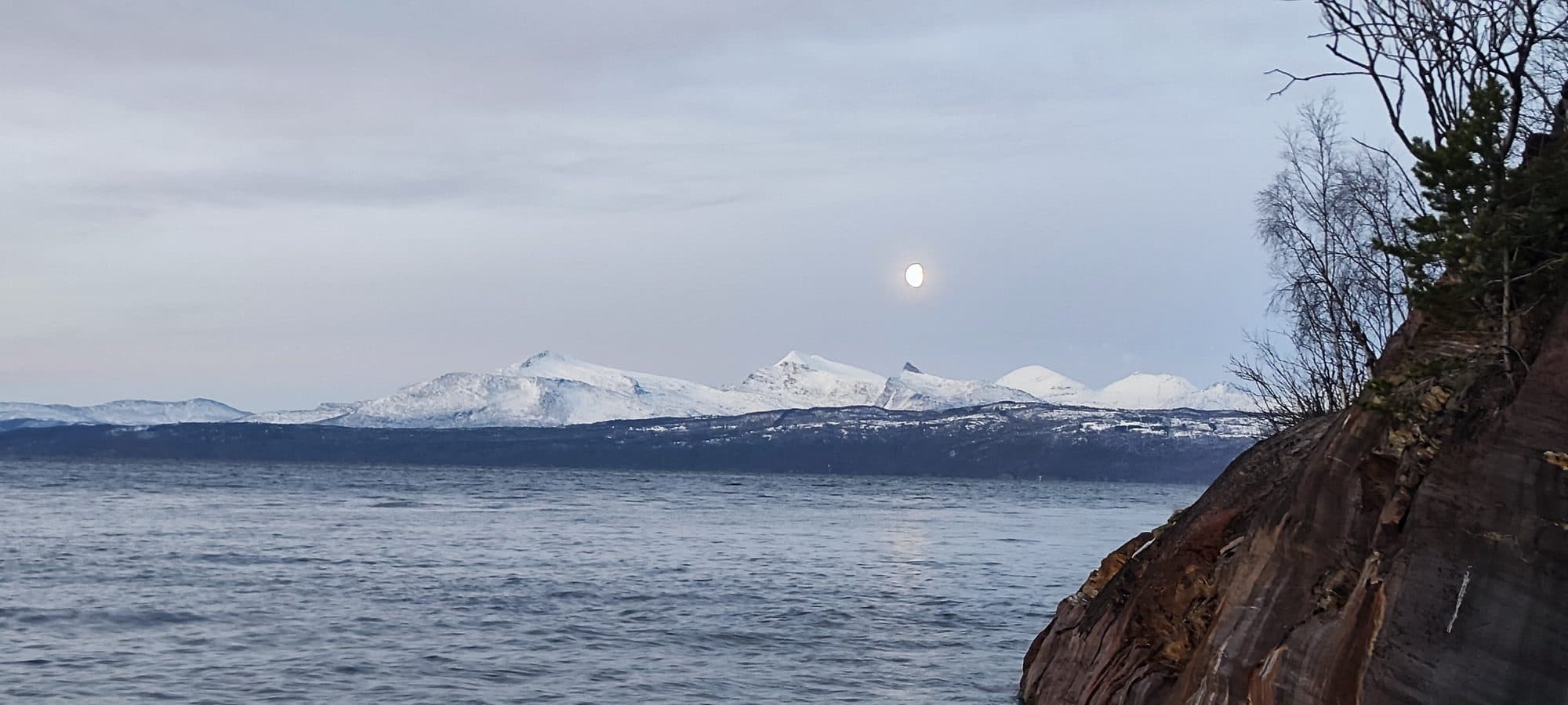 Der Vollmond steigt über schneebedeckten Gipfeln jenseits des Fjords auf, im Vordergrund eine schroffe Felskante.