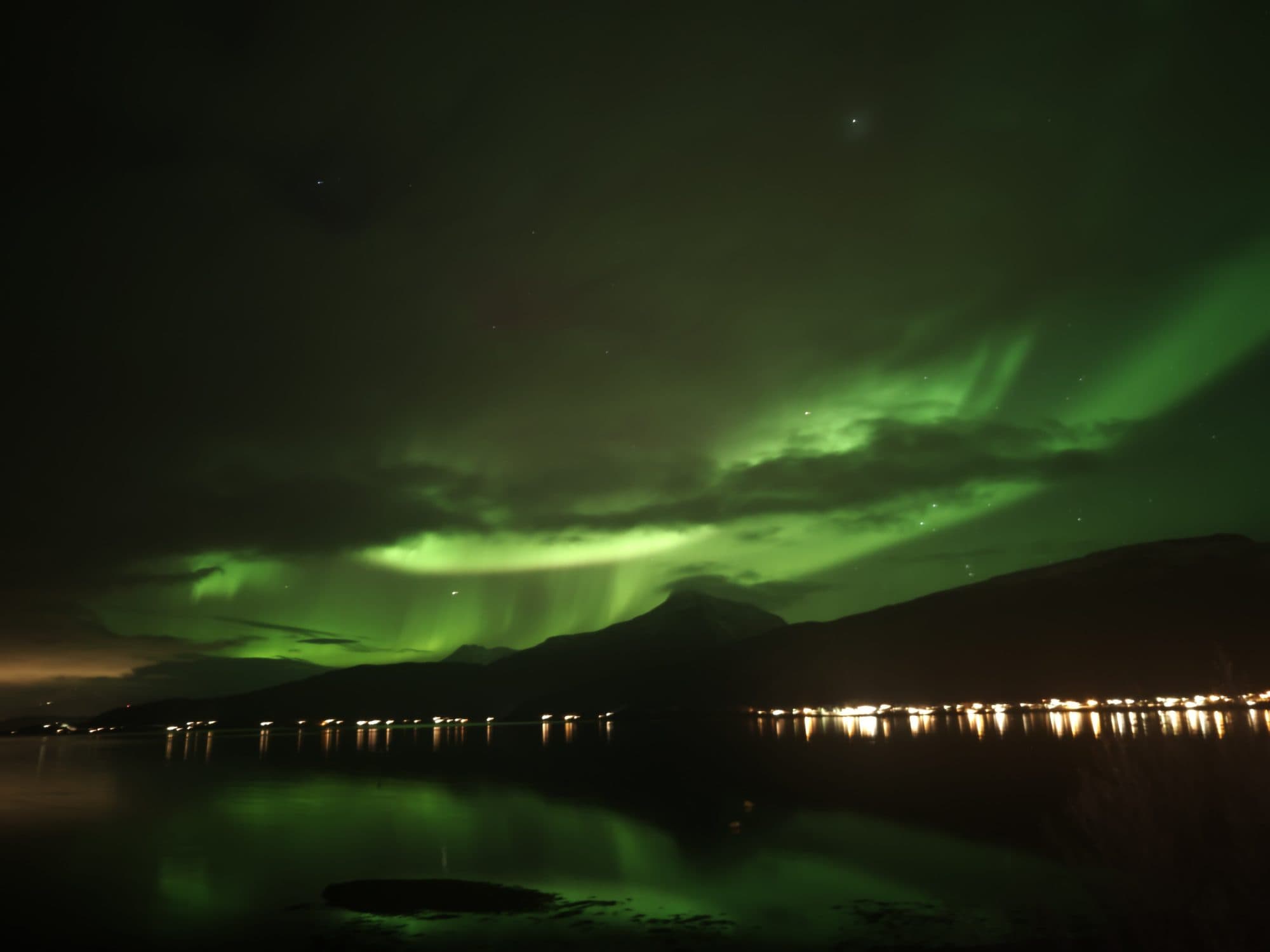 Ein leuchtend grüner Polarlichtbogen wölbt sich über den Fjord bei Ballangen und spiegelt sich im ruhigen Wasser.