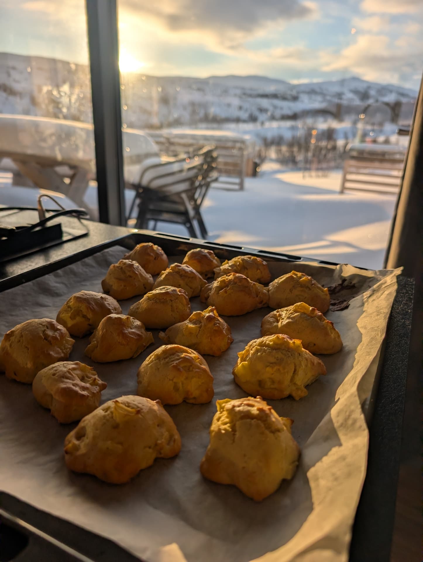 Nahaufnahme der goldbraunen Brötchen auf dem Backblech, das warme Licht der Morgensonne beleuchtet die Kruste, im Hintergrund sanft das Bergpanorama.