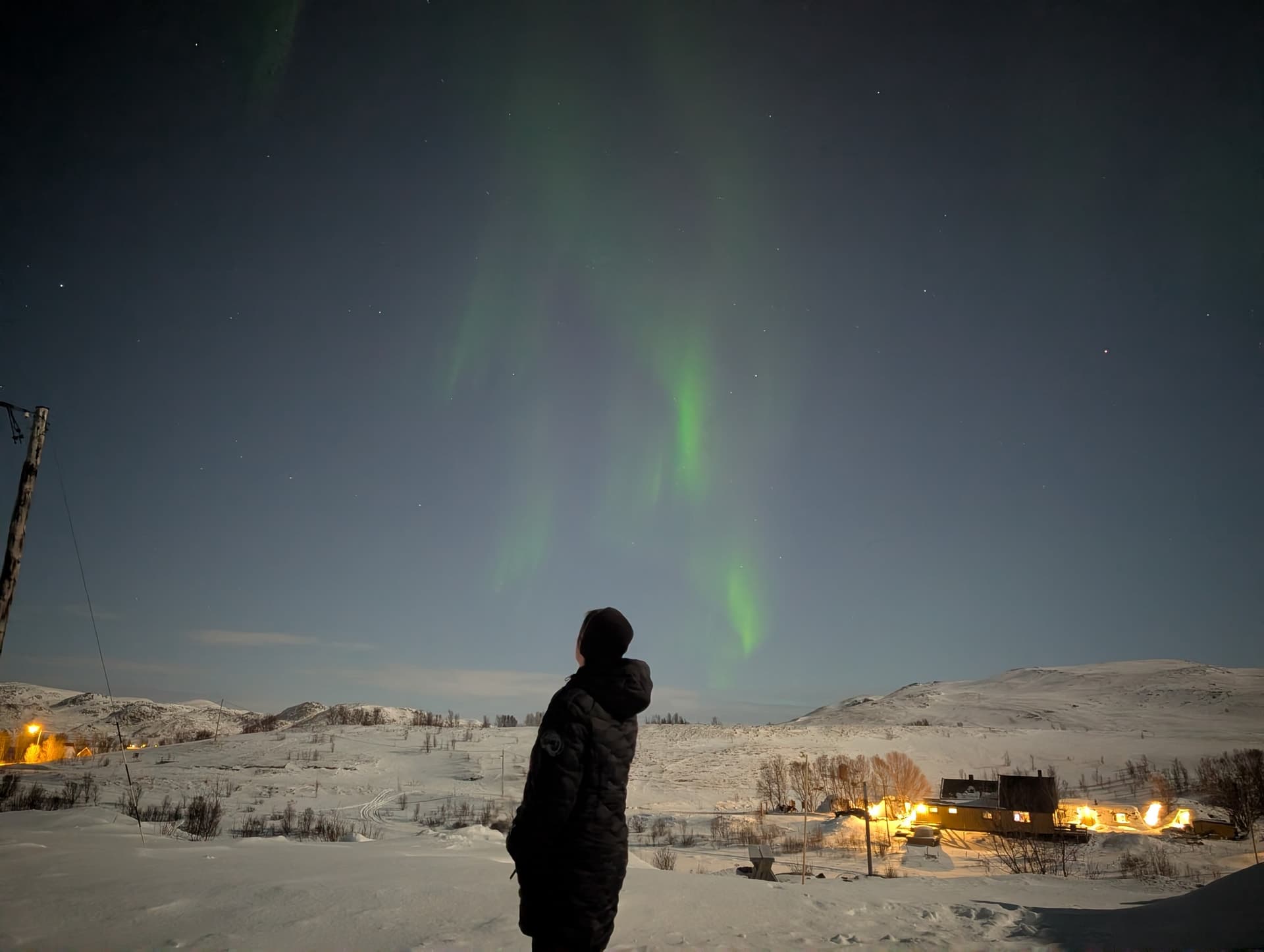 Eine Person bewundert bei Nacht die grünen Vorhänge des Nordlichts über einer verschneiten nordnorwegischen Landschaft mit leuchtenden Häusern im Hintergrund.