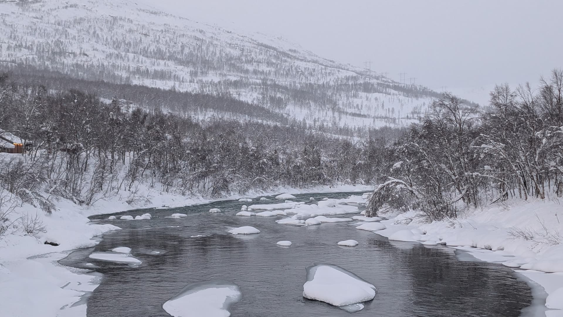 Dichter Schneefall zieht über einen winterlichen Flusslauf bei Bardu, während am linken Bildrand eine einzelne erleuchtete Hütte im Schneetreiben aufscheint.