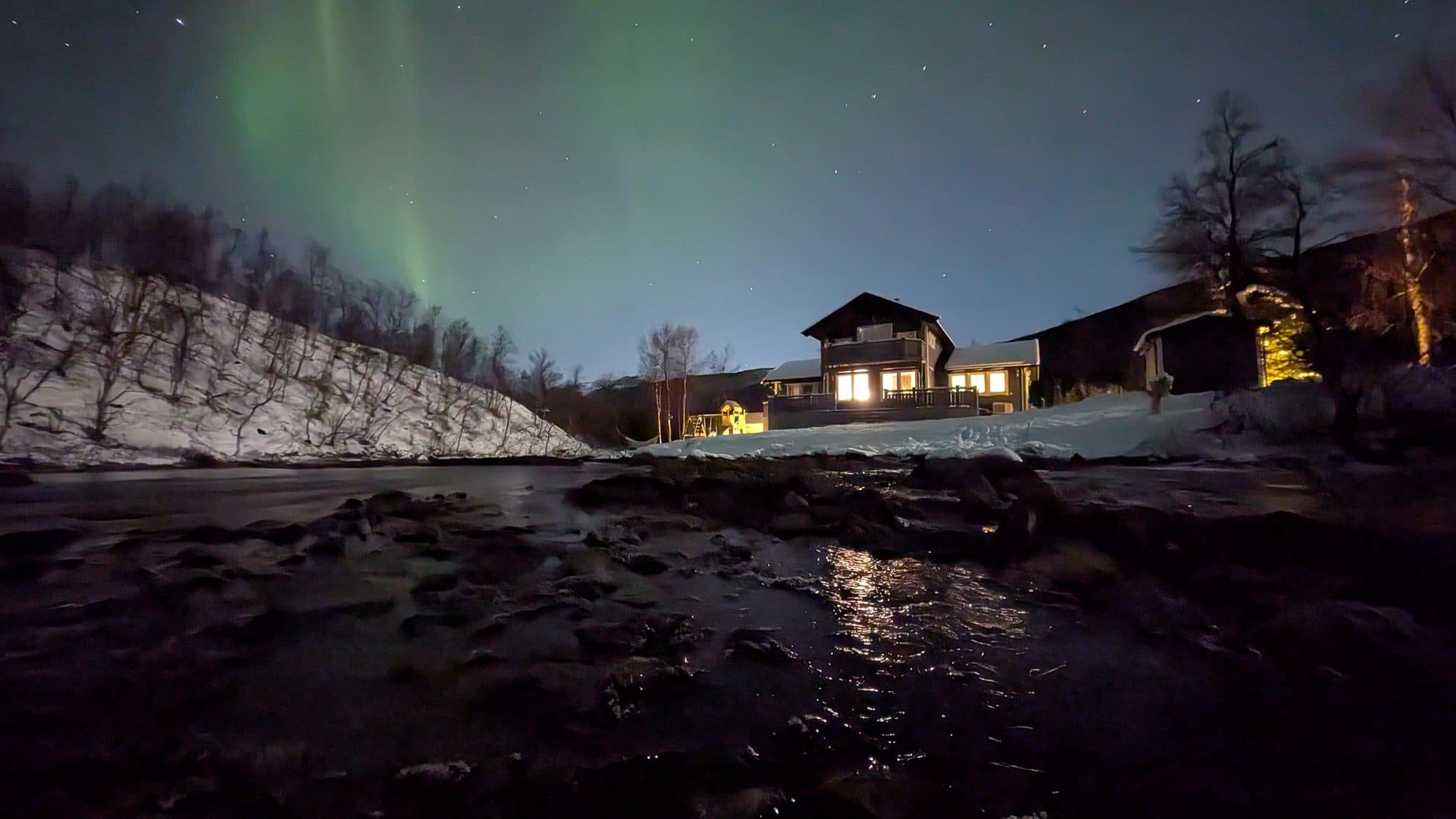 Über dem warm erleuchteten Ferienhaus bei Narvik tanzt ein grünes Polarlichtband am Sternenhimmel.