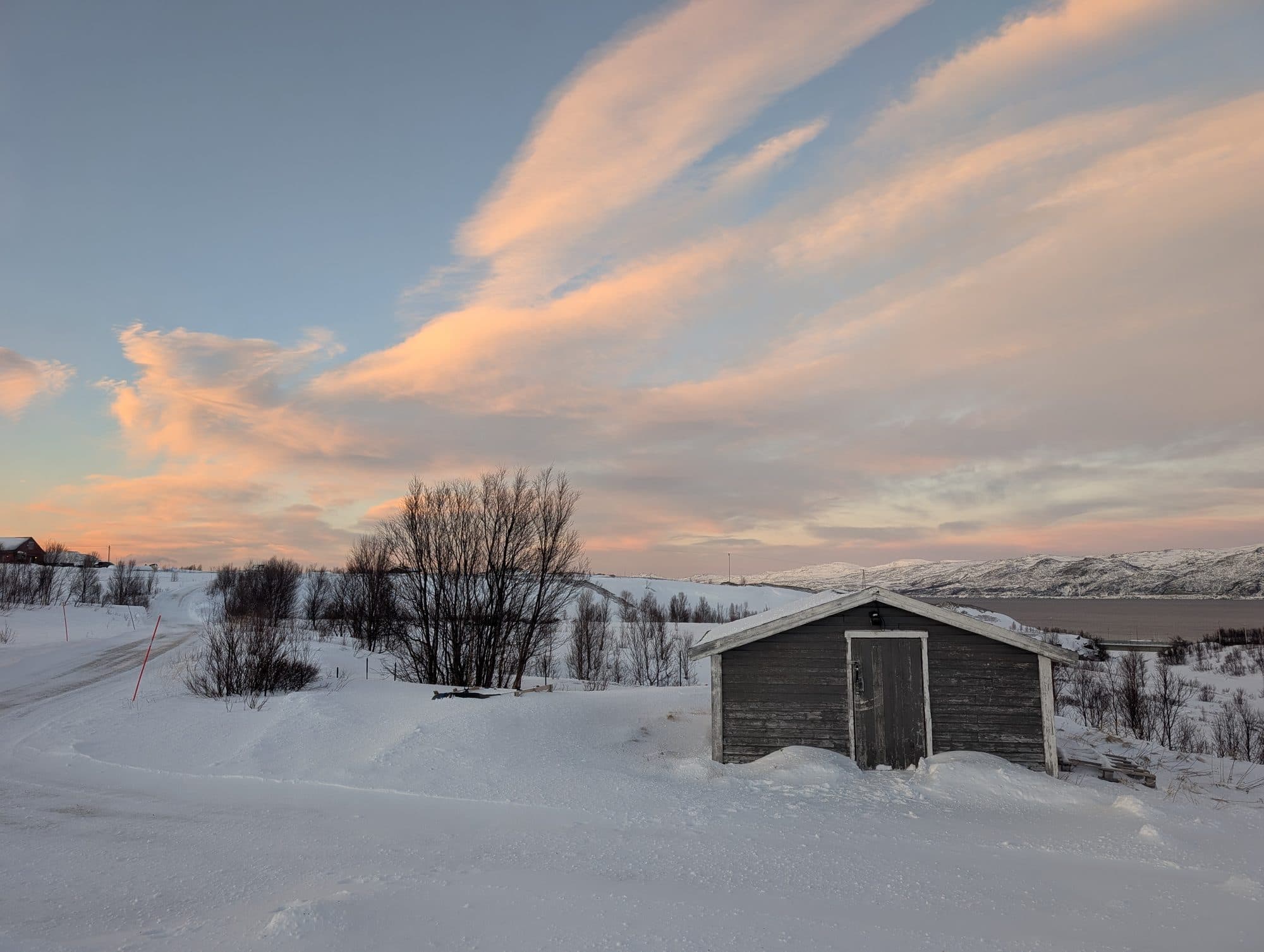 Verfallener Holzschuppen vor dramatischem Pastellhimmel mit rosa Wolken und Fjord.