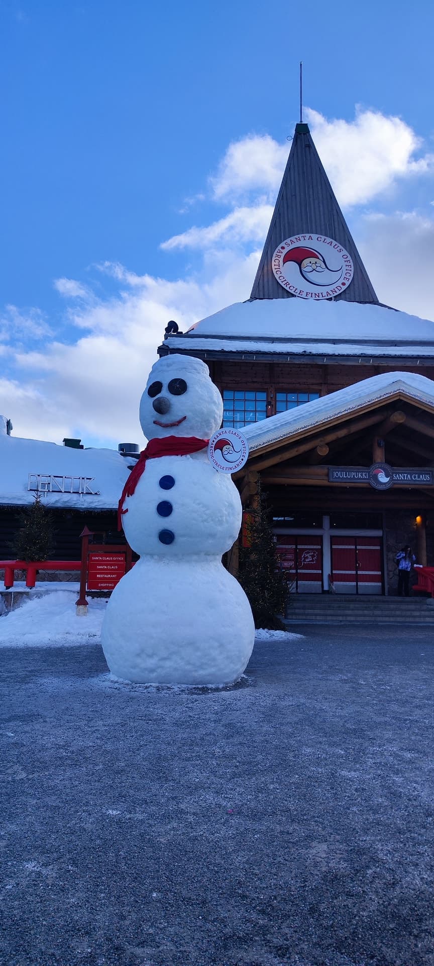 Ein großer Schneemann mit rotem Schal und blauen Knöpfen steht vor dem spitzen Turm des Santa Claus Office am Polarkreis in Rovaniemi, dahinter strahlend blauer Himmel.